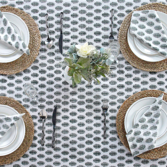 Table setting with a leaf patterned tablecloth, white plates, and woven placemats.