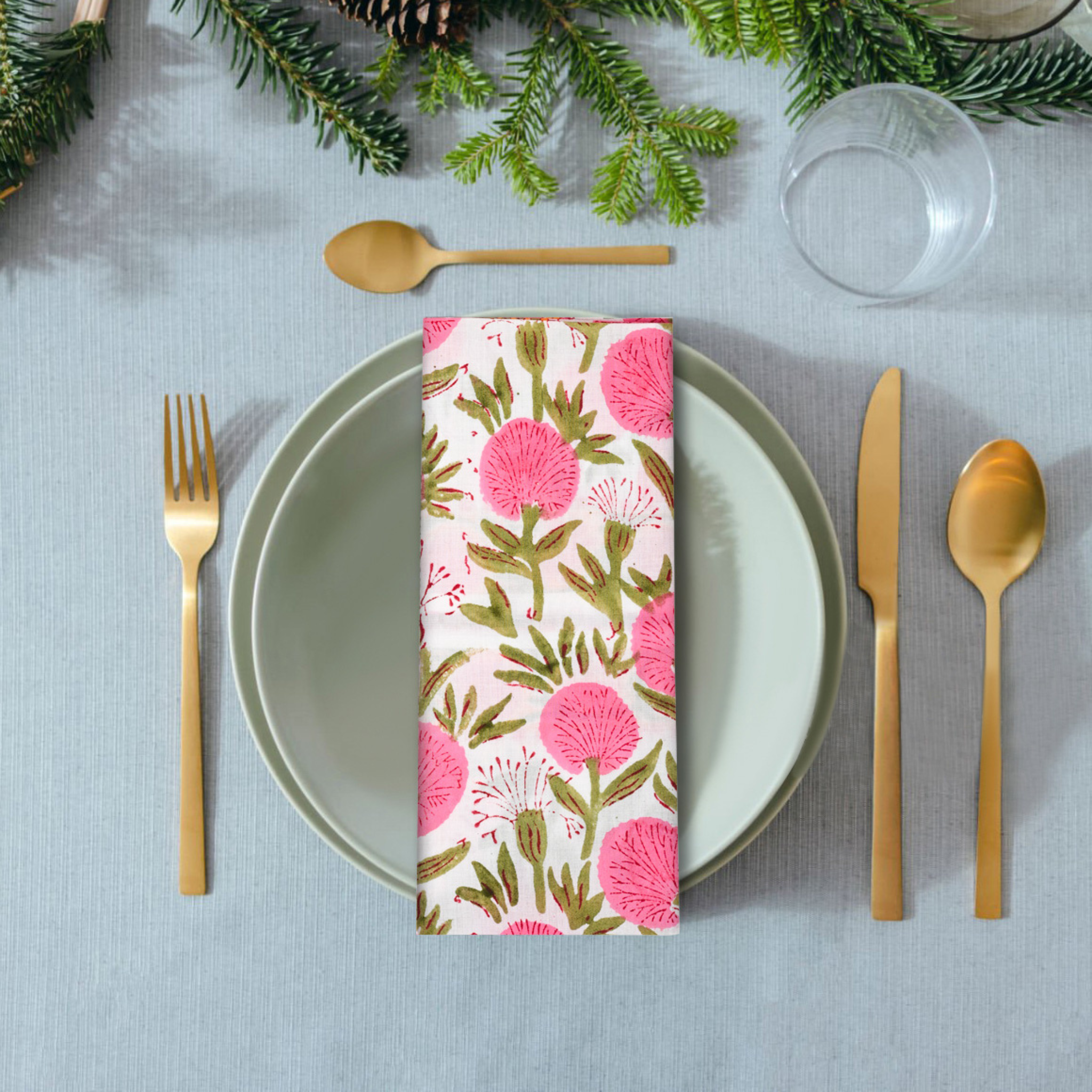 Table setting with floral-patterned napkin, gold cutlery, and greenery on a light blue tablecloth.