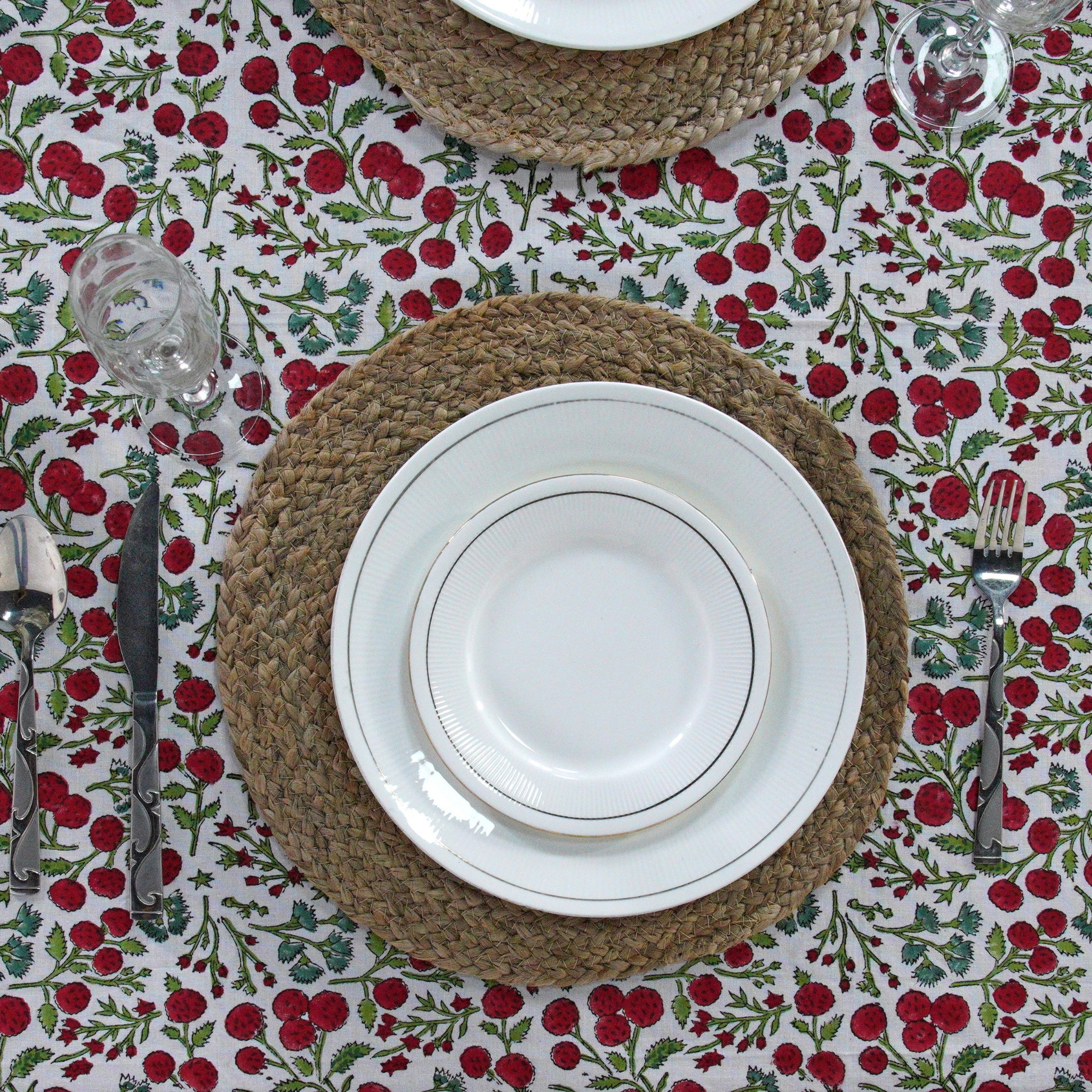 Table setting with white plates on woven placemats over a red floral tablecloth.