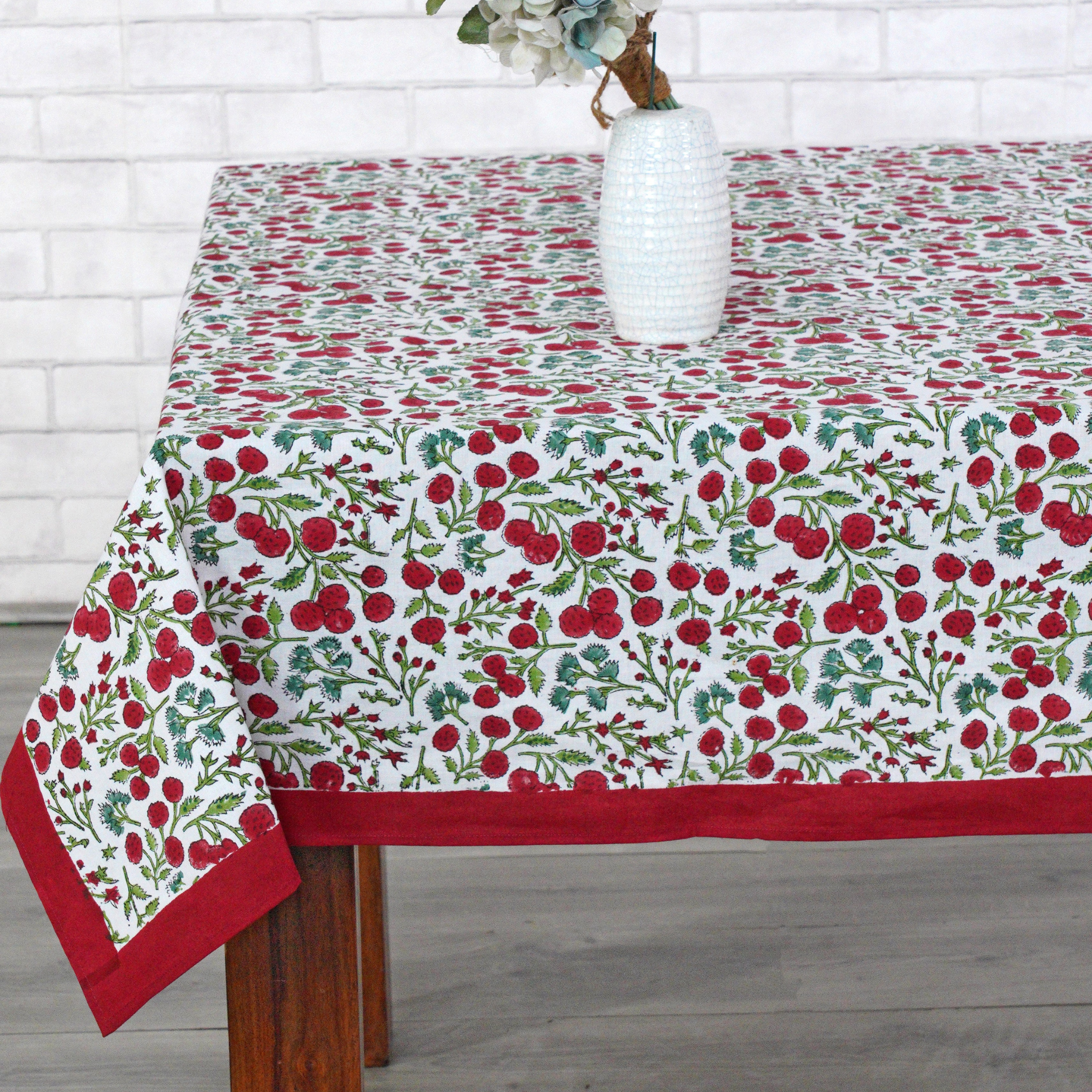 Tablecloth with red floral pattern on a wooden table against a white brick wall.