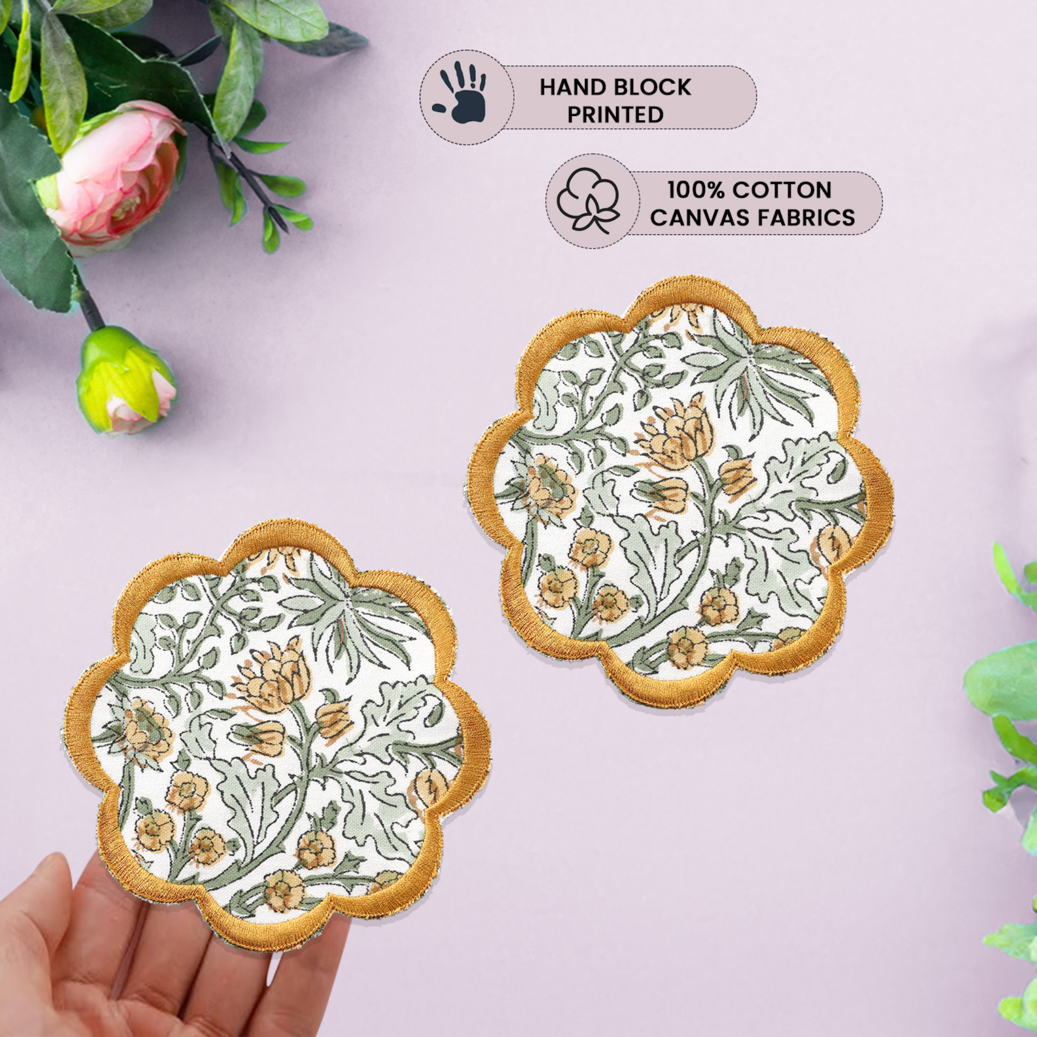 Two floral coasters with gold edges on a light pink background, held by a hand.