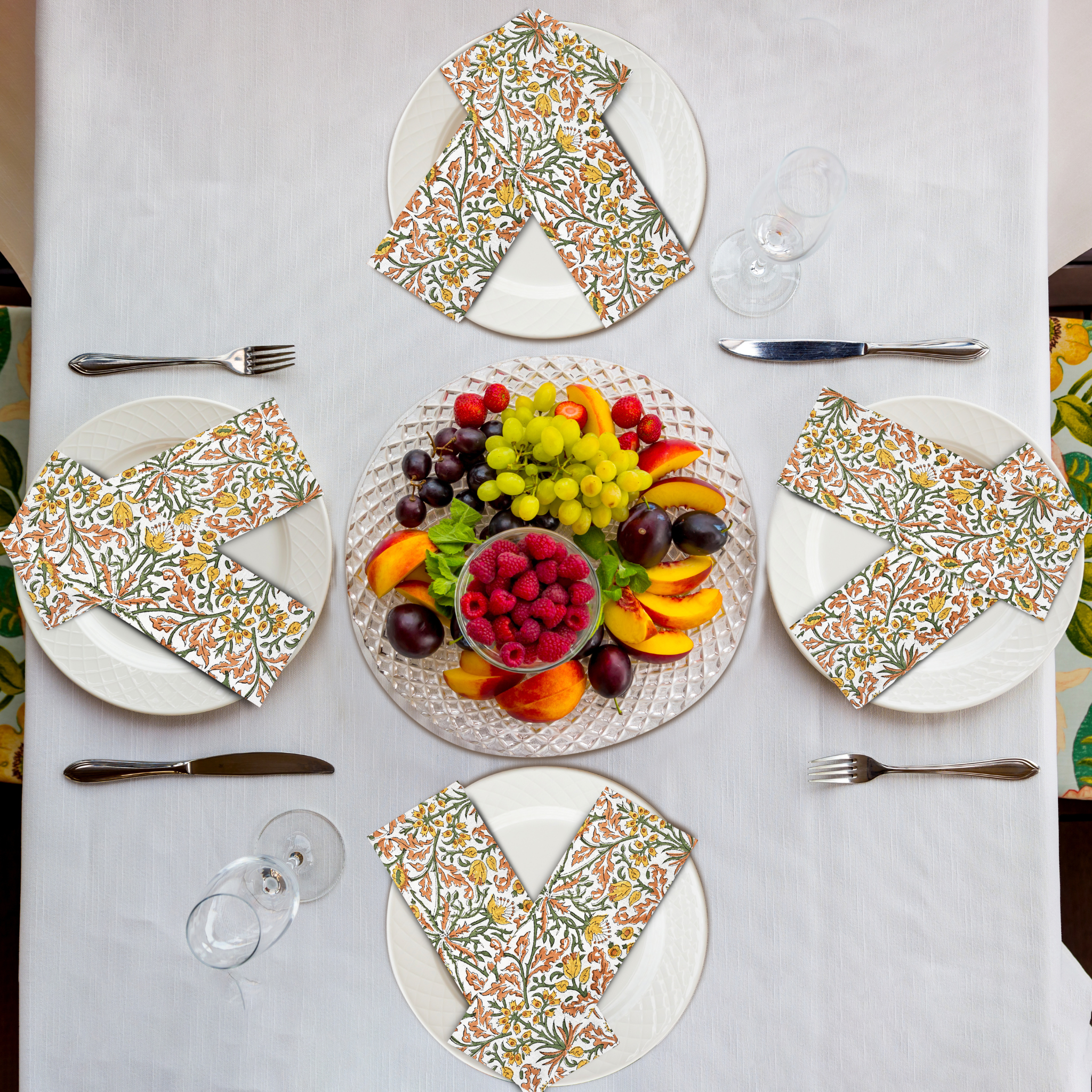 Table setting with floral napkins, fruit platter, and cutlery on a white tablecloth.