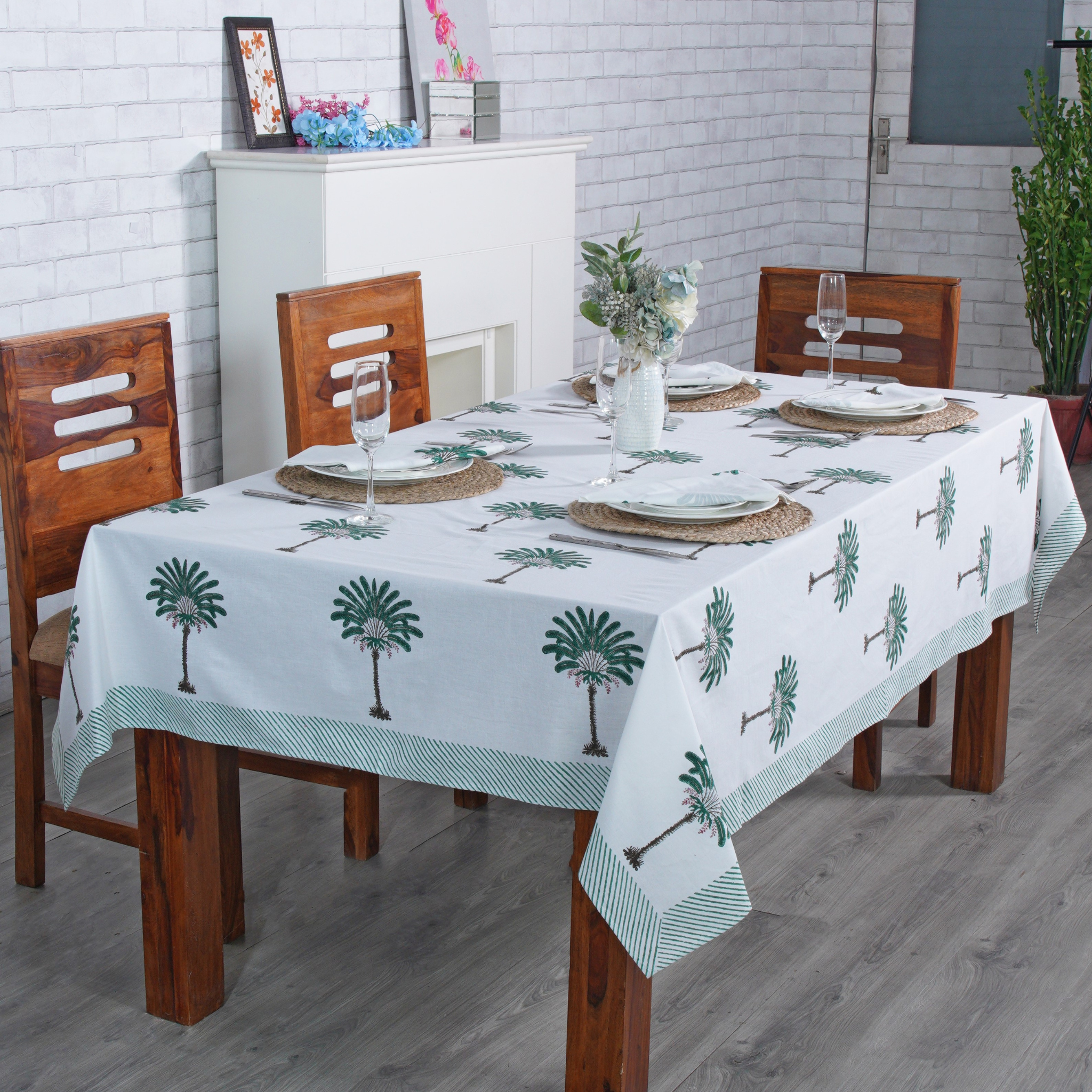 Dining table set with a palm tree patterned tablecloth in a room with wooden chairs and a white brick wall.