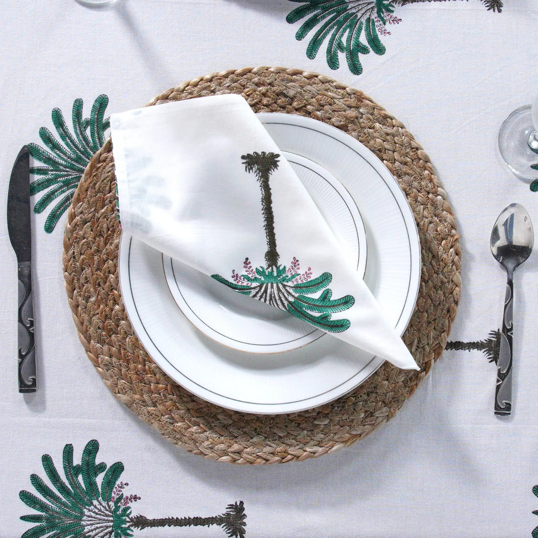 Table setting with a palm tree-themed napkin on a white tablecloth with green leaf patterns.