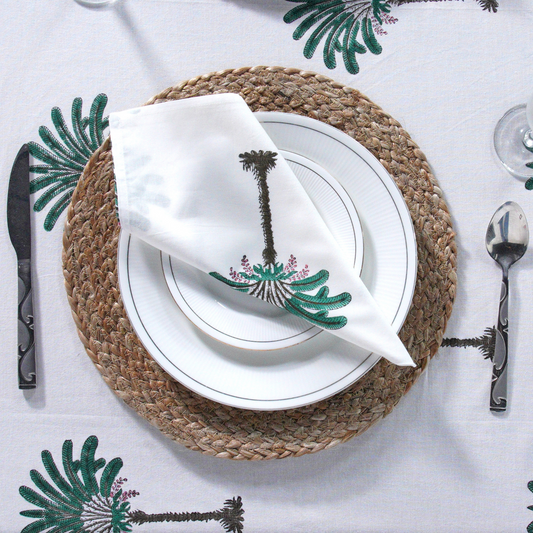 Table setting with a palm tree-themed napkin on a white tablecloth with green leaf patterns.