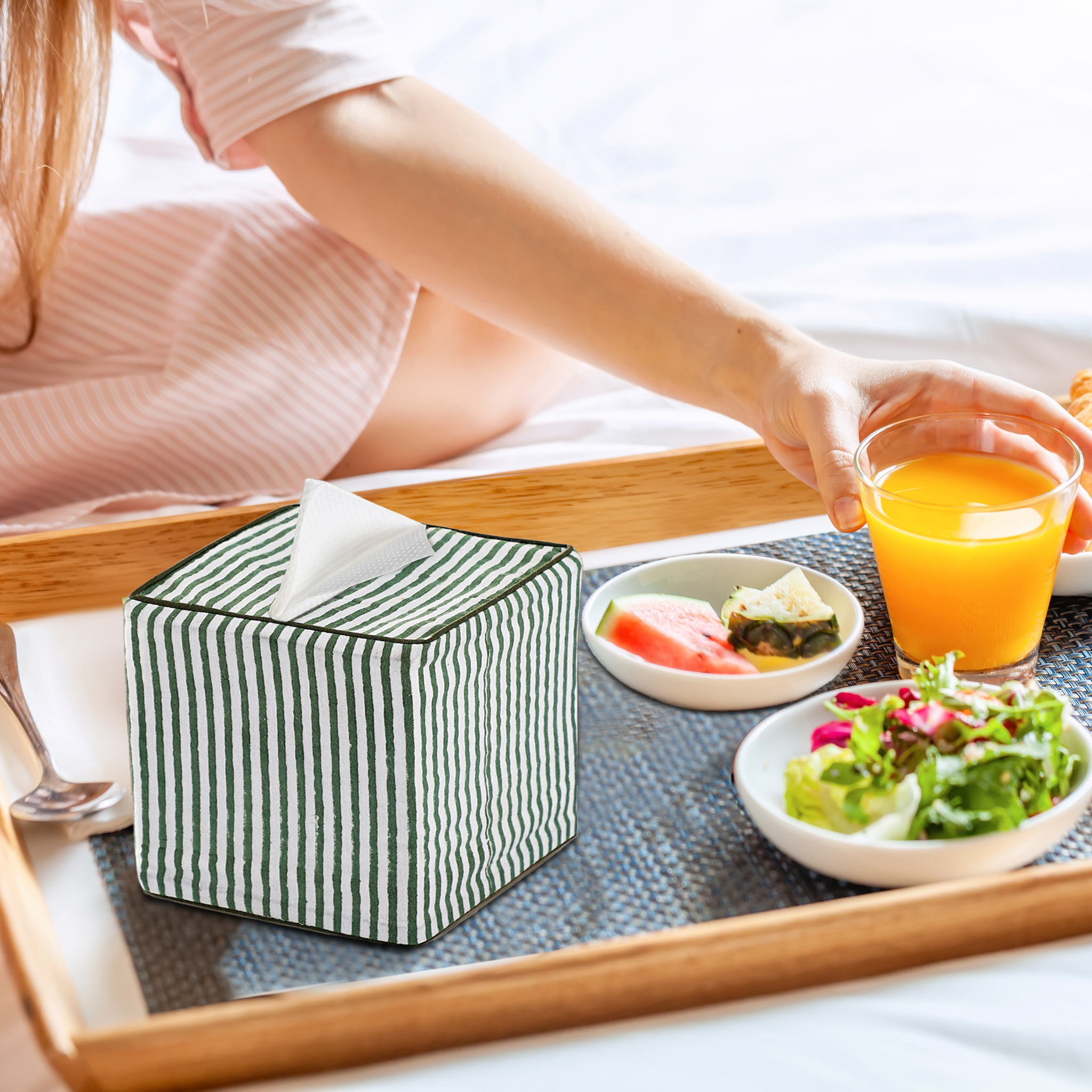 Person holding a glass of orange juice next to a striped tissue box on a tray with food.
