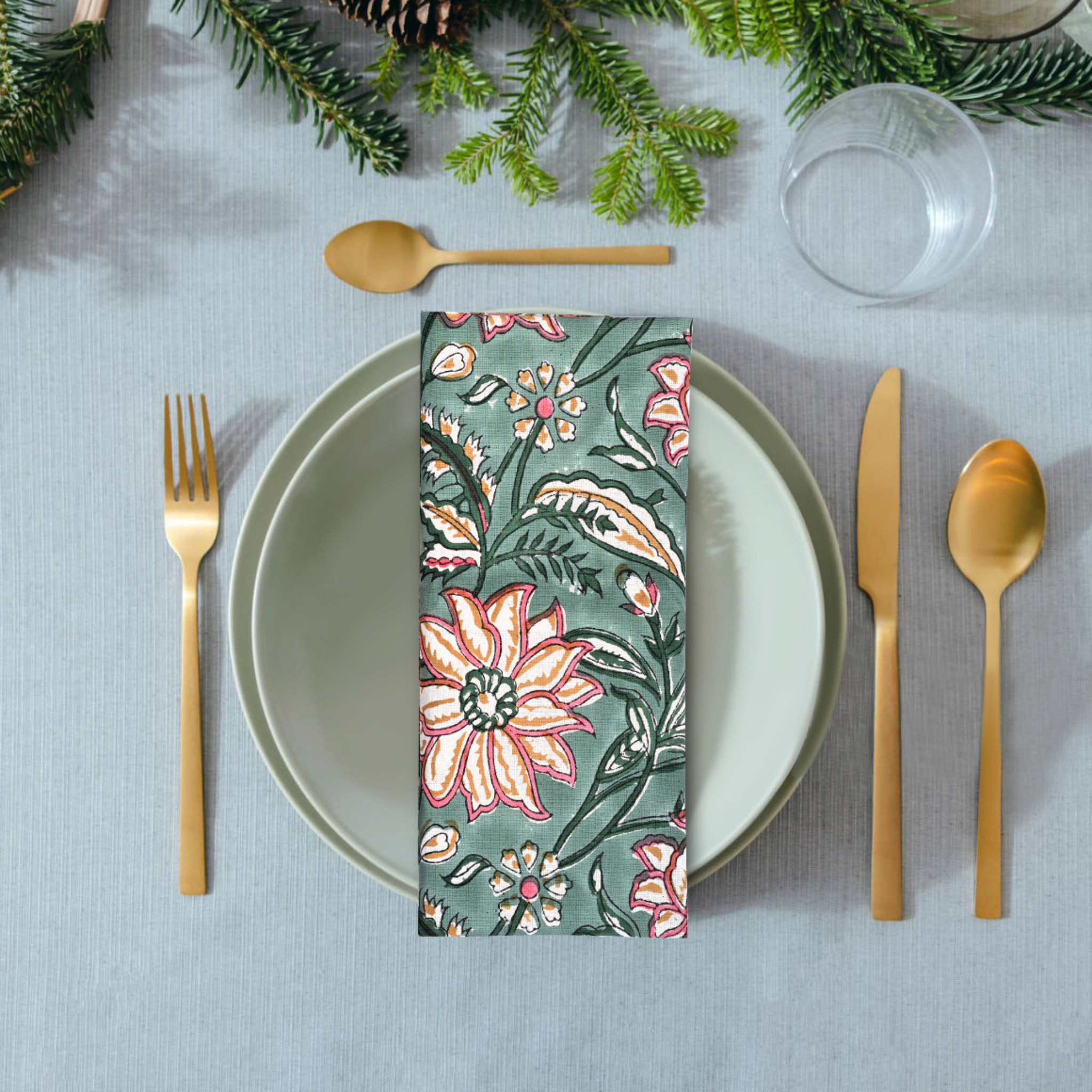 Table setting with floral napkin, gold cutlery, and greenery on a gray tablecloth.