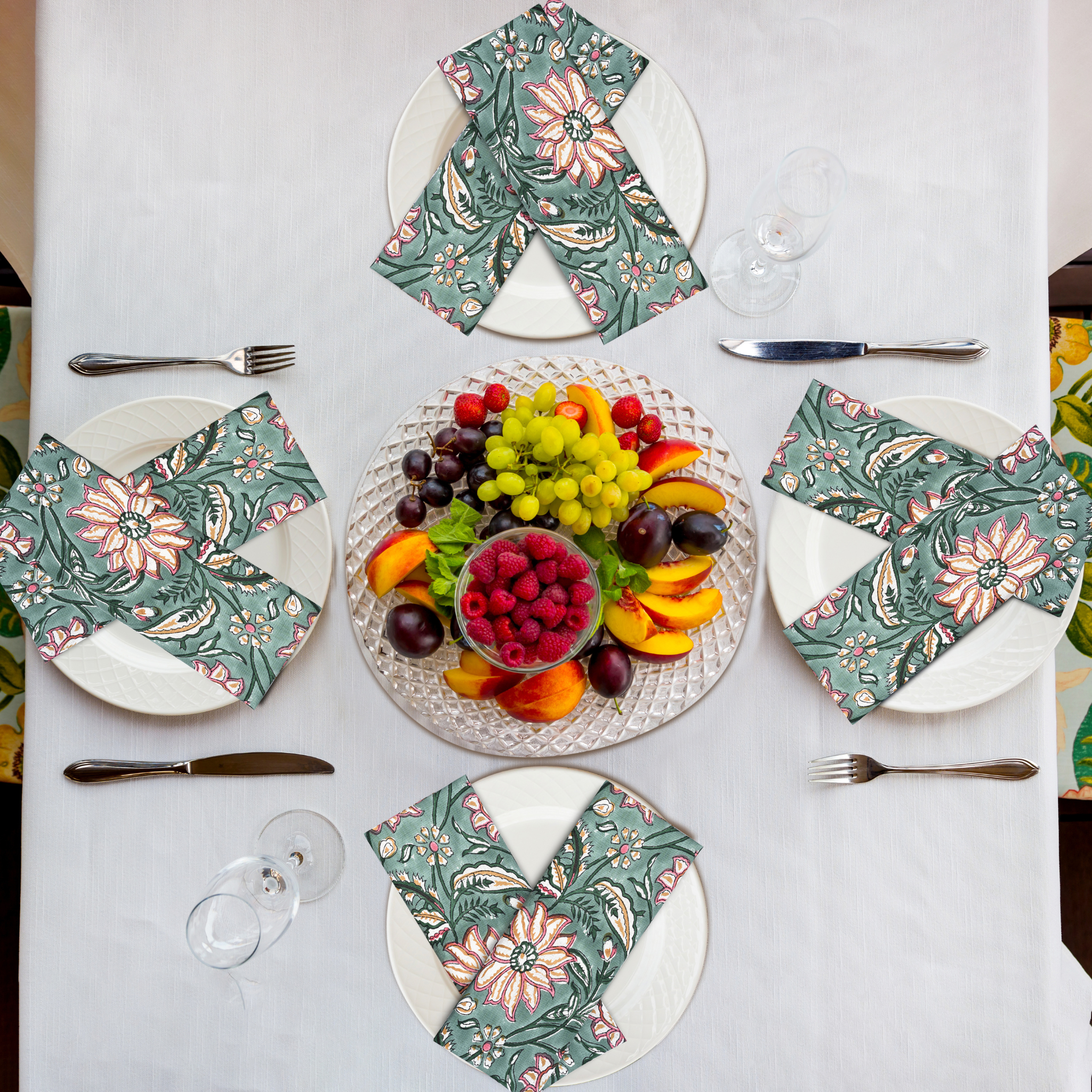 Table setting with floral napkins, fruit platter, and cutlery on a white tablecloth.
