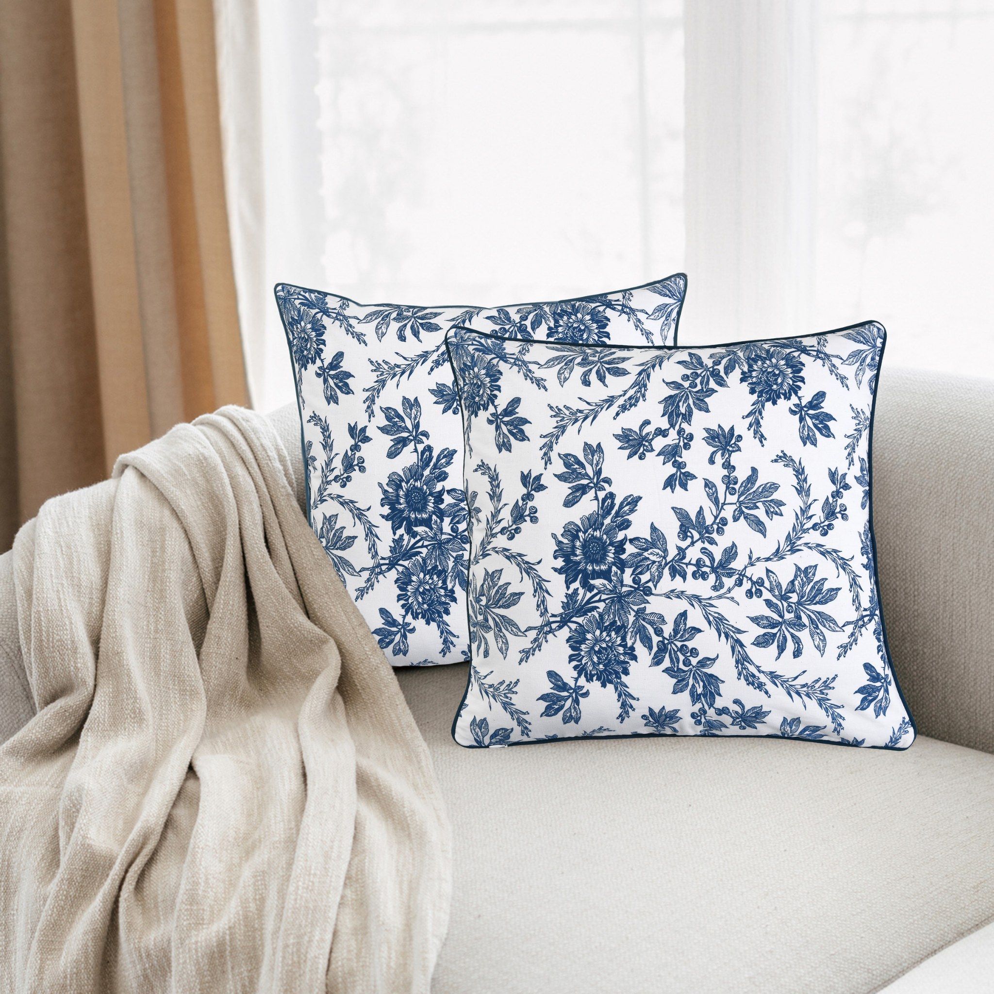 Two blue floral patterned pillows on a beige sofa with a neutral curtain in the background.