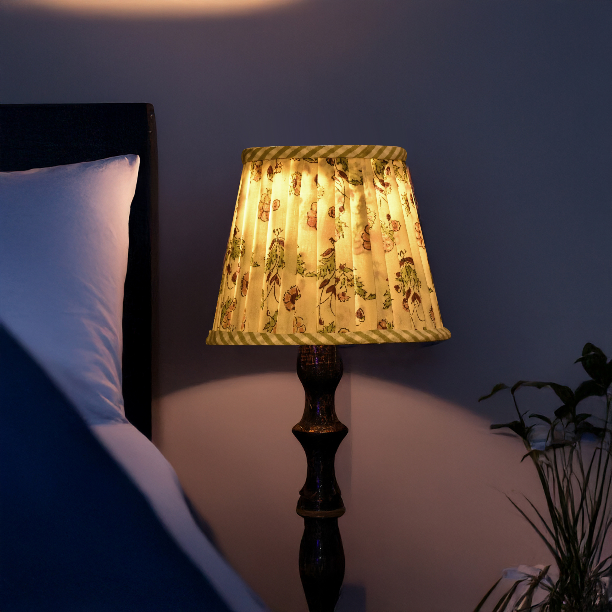 Lamp with patterned shade on a bedside table next to a bed in a dimly lit room.