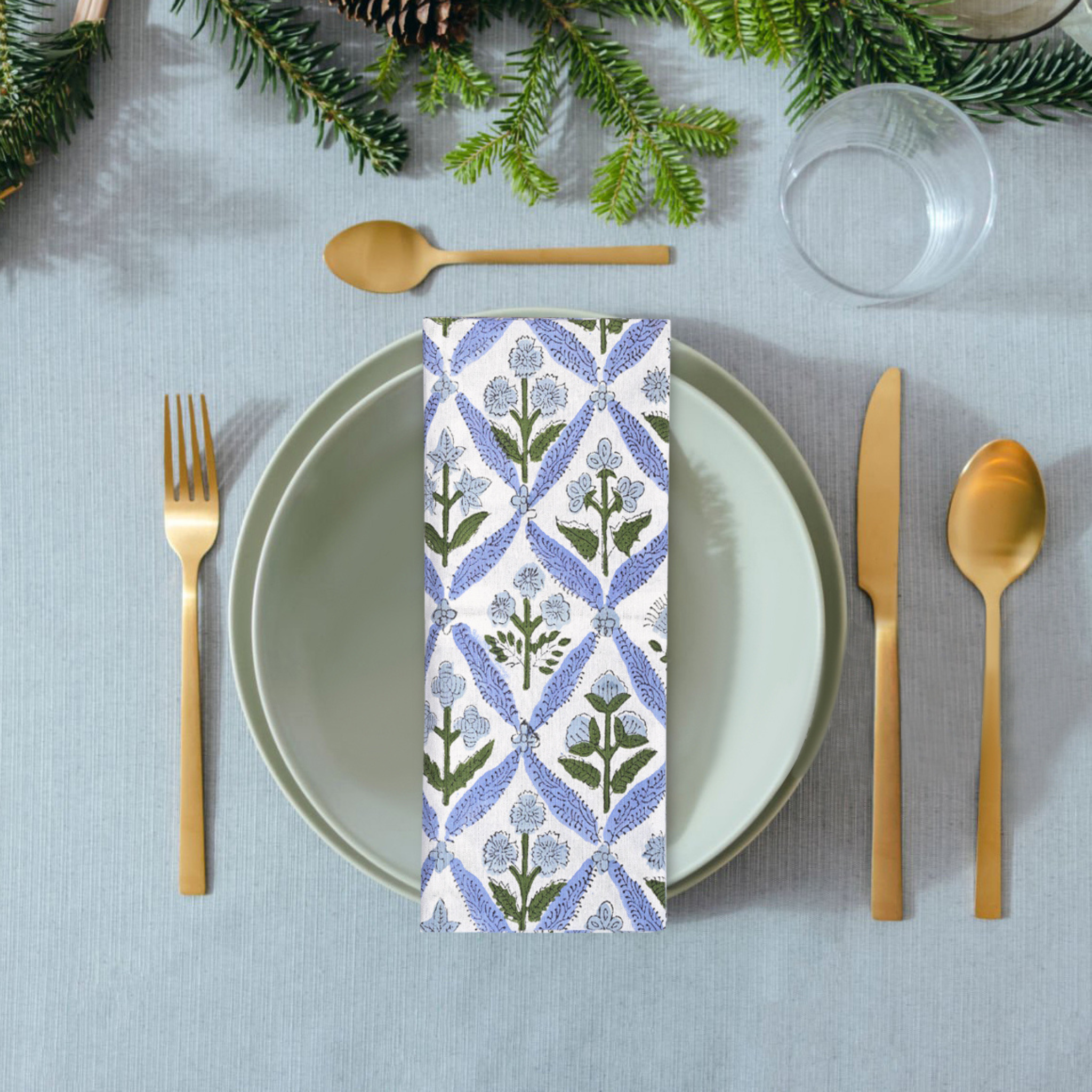 Table setting with a floral-patterned napkin, gold cutlery, and greenery on a gray tablecloth.