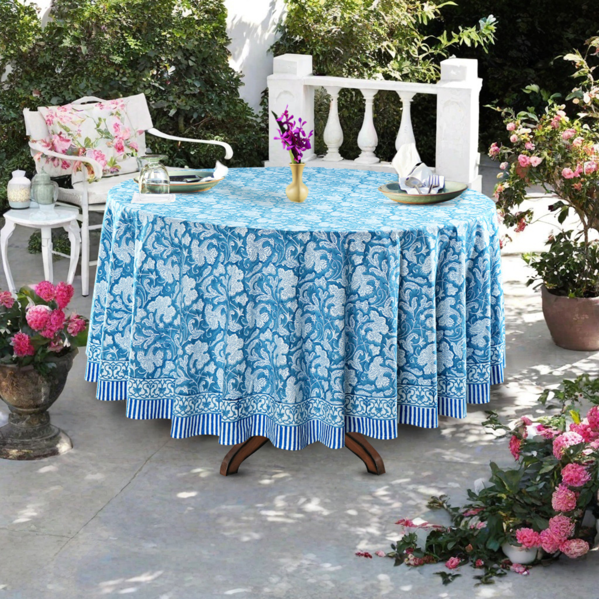Outdoor setting with a table covered by a blue floral-patterned tablecloth, surrounded by potted plants and chairs.