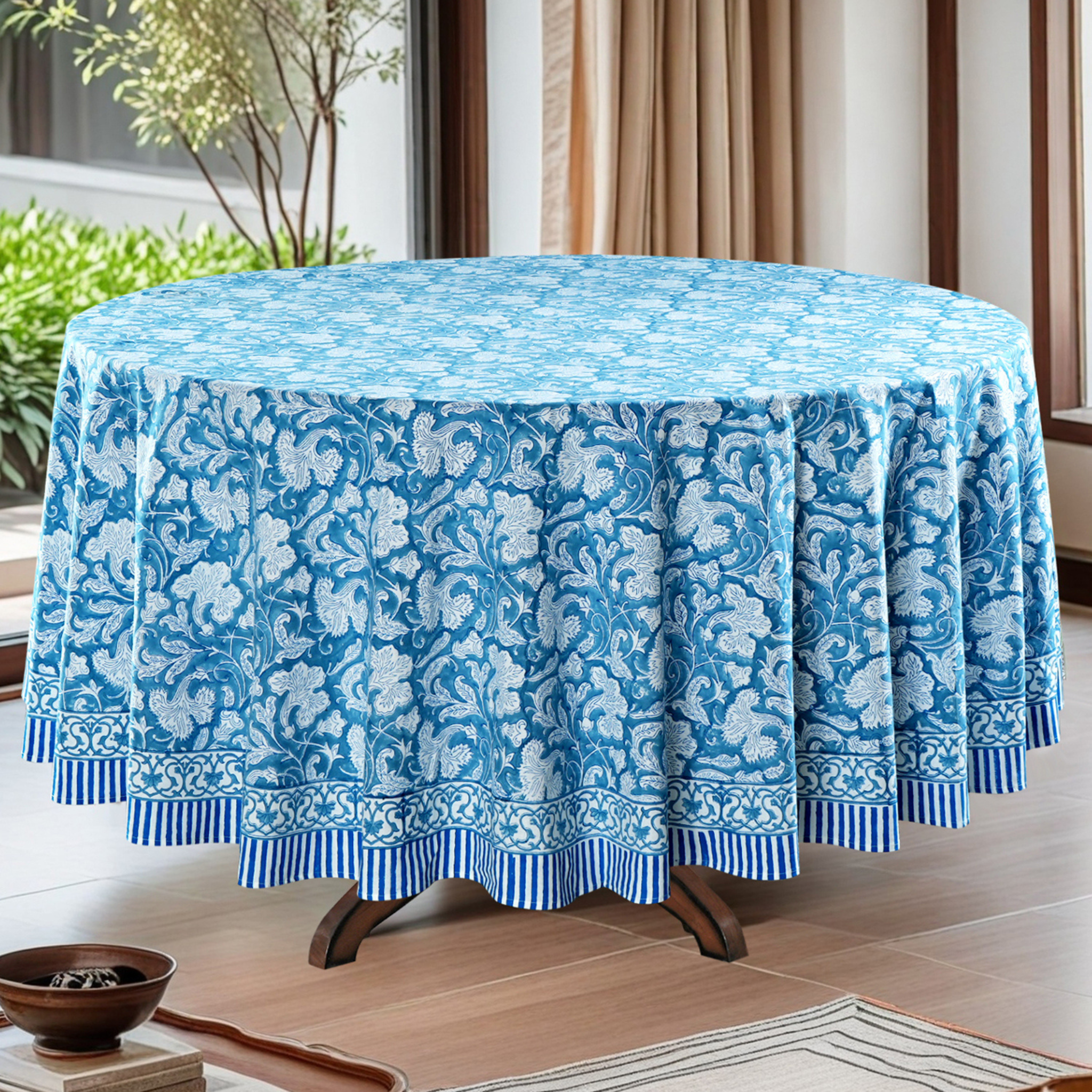 Round table with a blue floral patterned tablecloth in a room with plants and curtains.