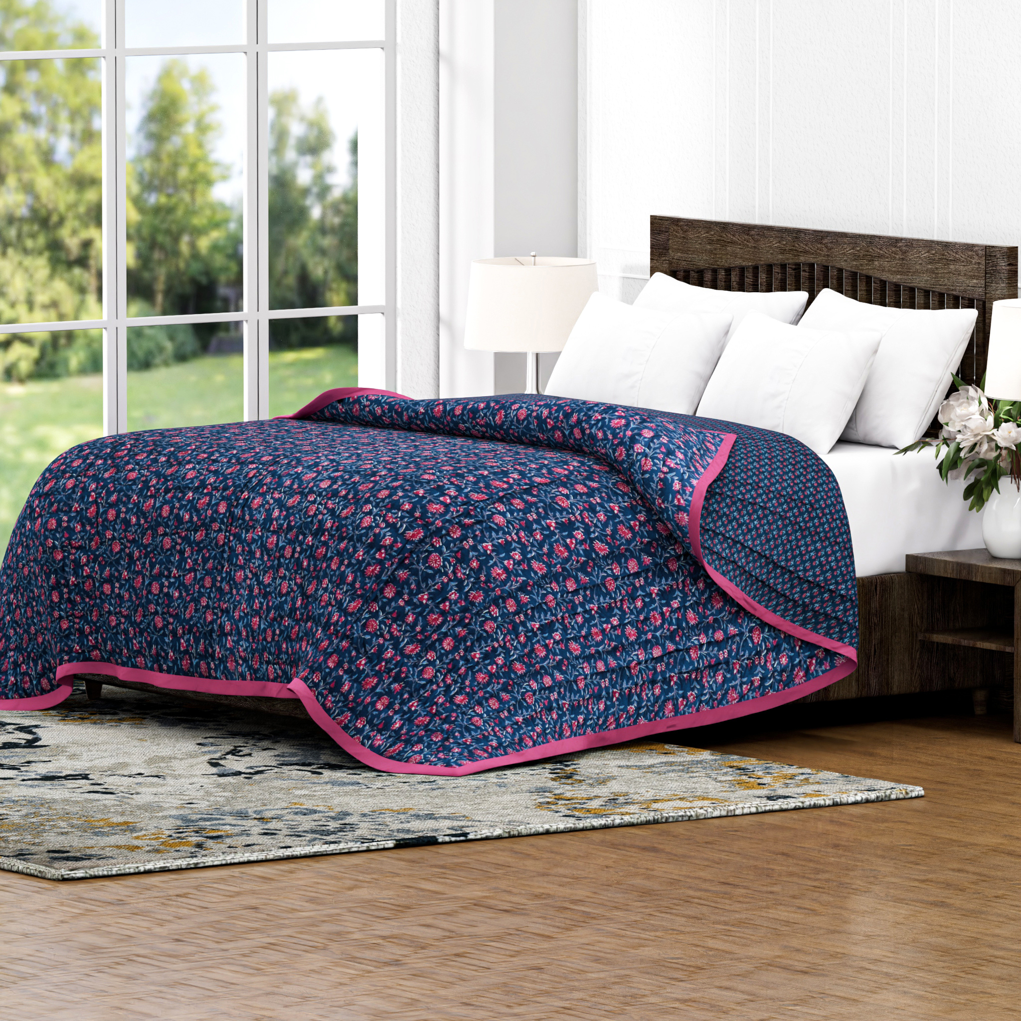 Blue floral quilt with pink trim on a bed in a room with large windows.