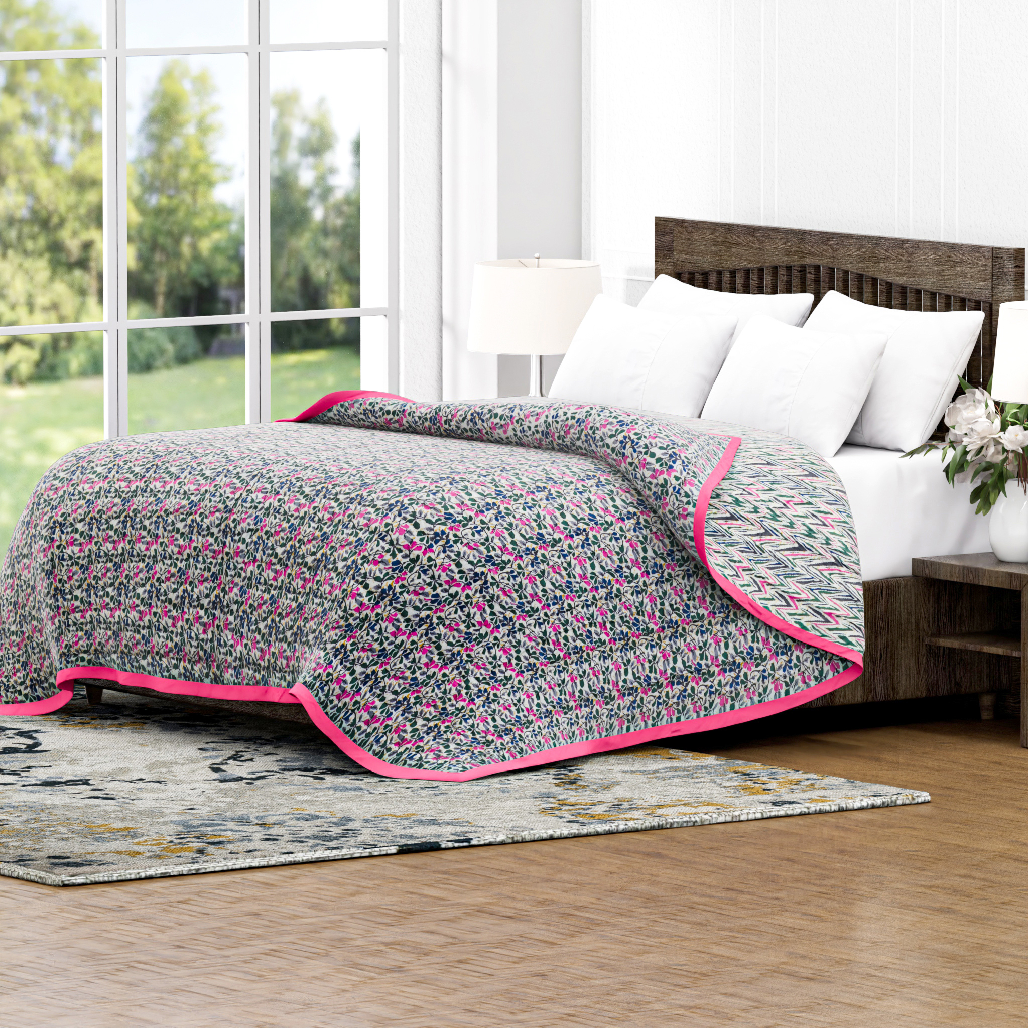 Floral quilt with pink border on a bed in a bright room with large windows.