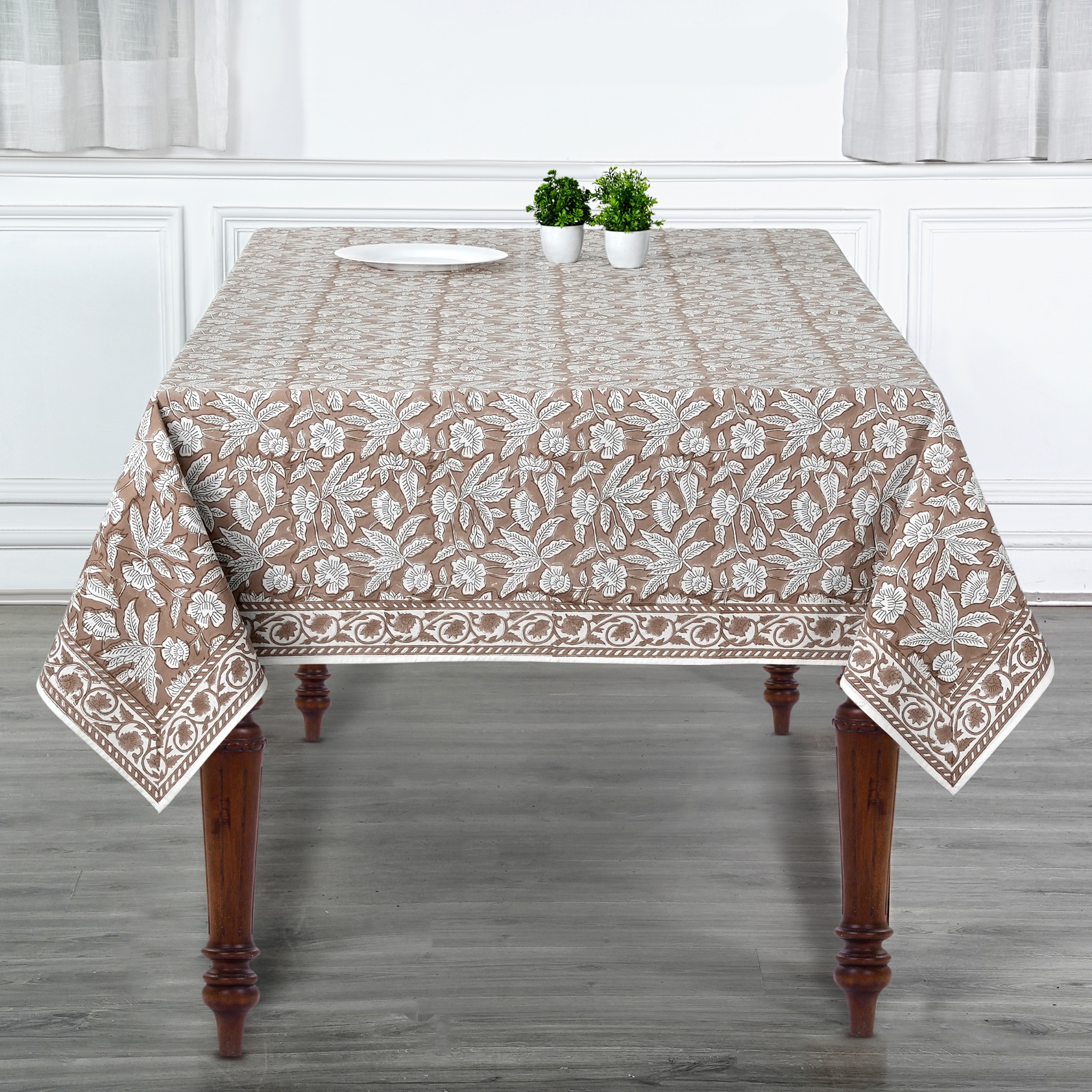 Tablecloth with floral pattern on a wooden table in a room with white walls and gray floor.