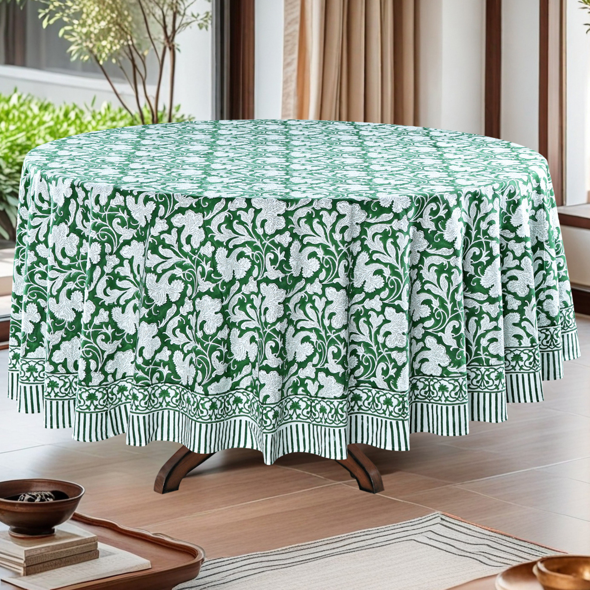 Round table with a green and white floral patterned tablecloth in a room with plants and curtains.