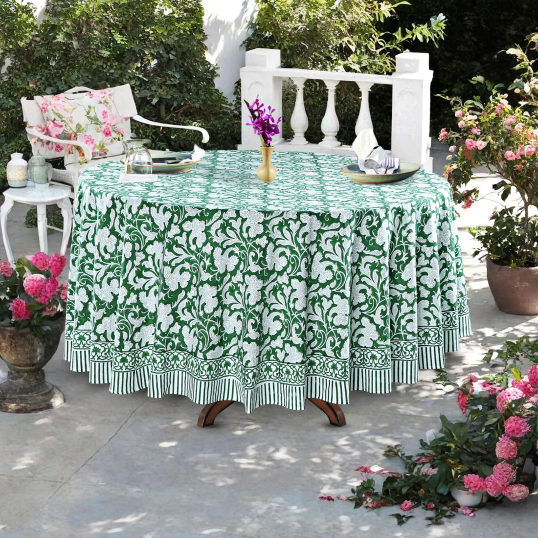 Table with a green and white floral tablecloth outdoors, surrounded by potted plants.