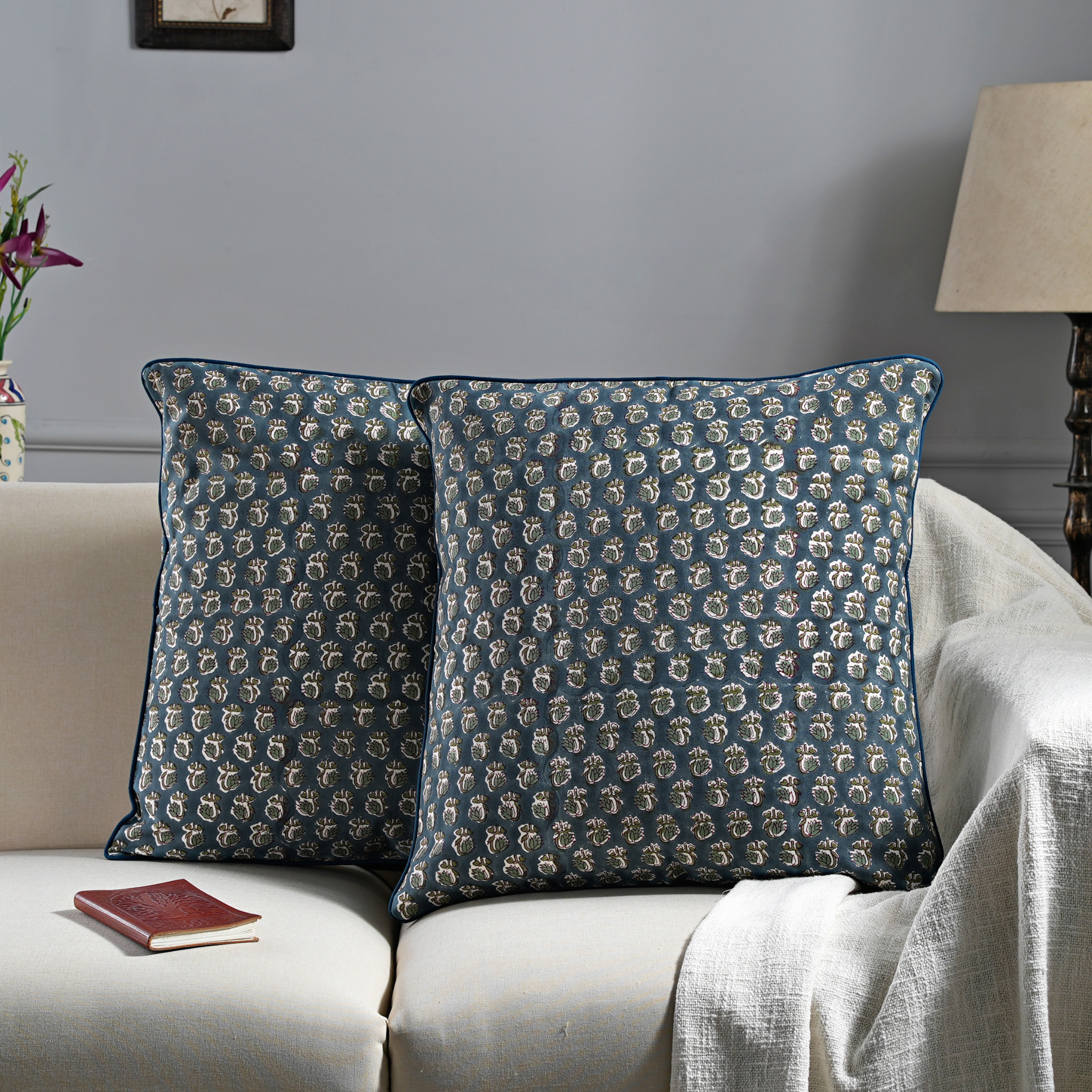 Two patterned cushions on a sofa with a lamp and flowers in the background.