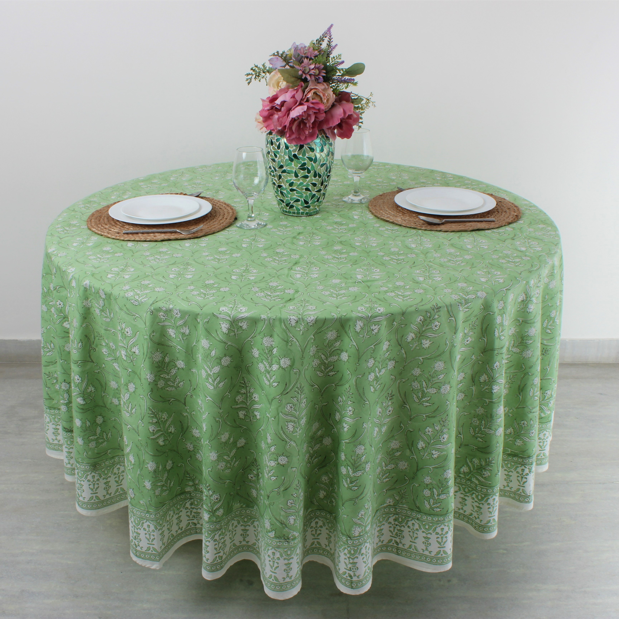 Round table with green floral tablecloth, plates, glasses, and a vase of flowers.