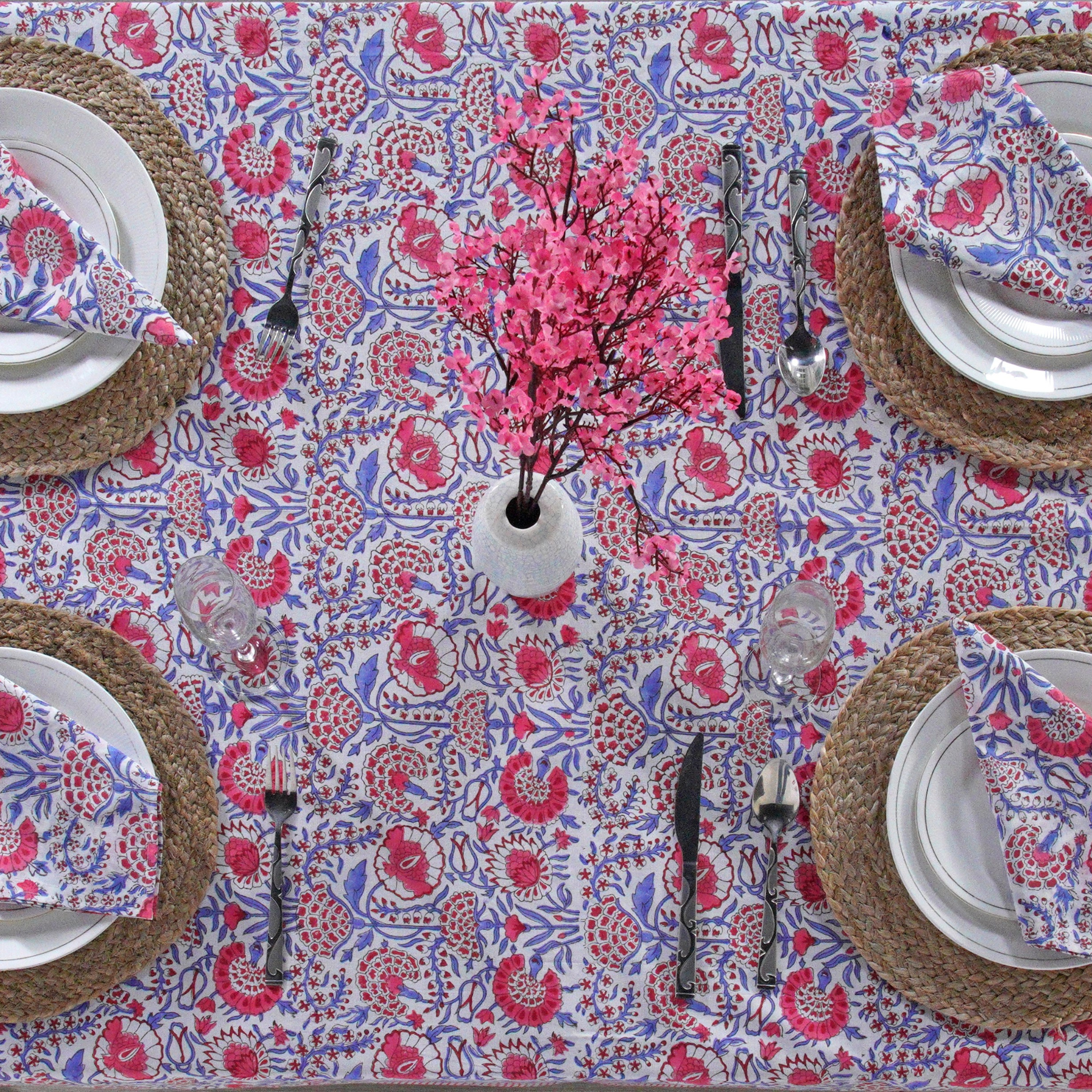 Decorative table setting with a floral patterned tablecloth, plates, cutlery, and a vase with pink flowers.
