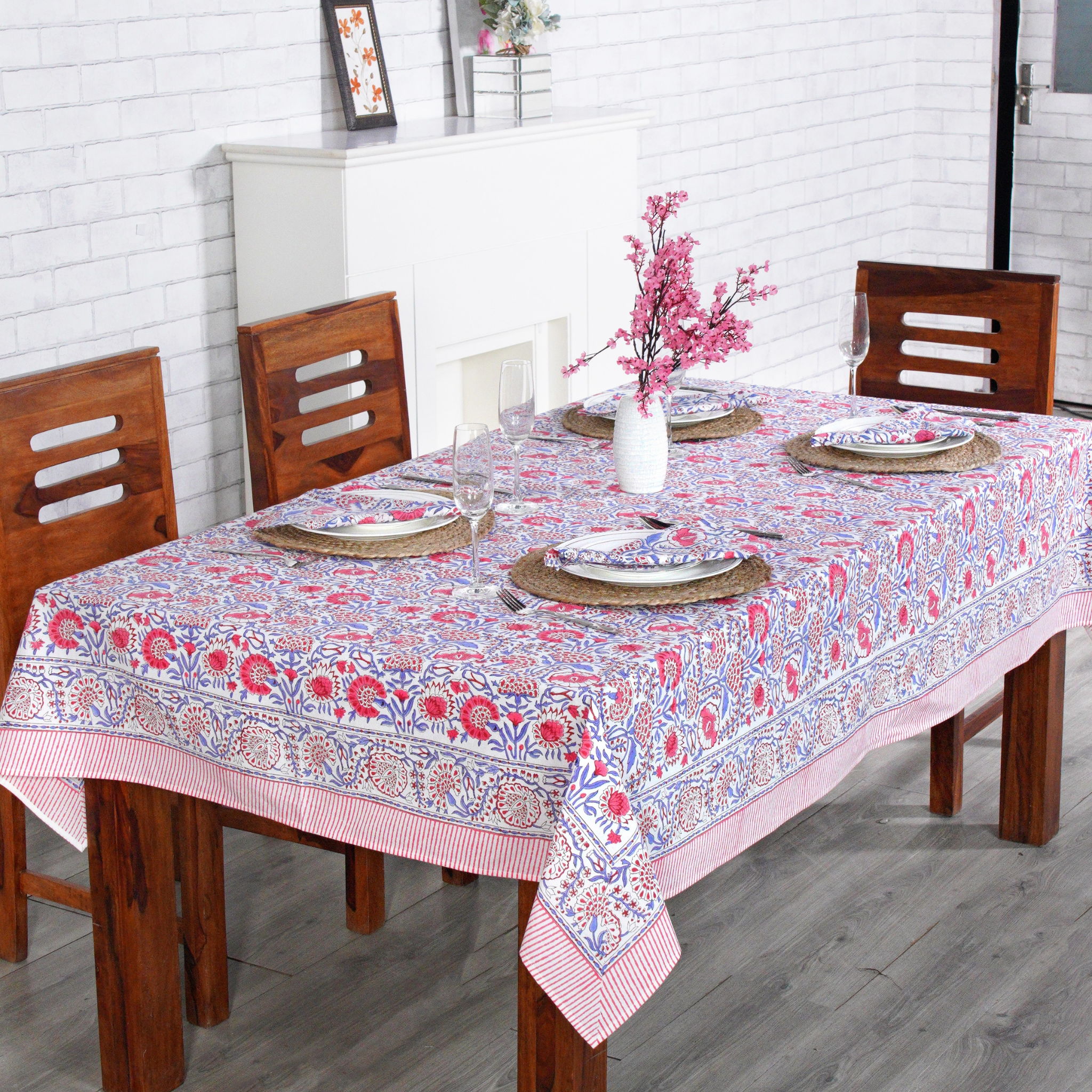 Dining table set with a floral tablecloth, plates, and chairs in a room with white brick walls.