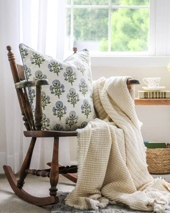 Wooden rocking chair with a patterned pillow and white blanket in a bright room.