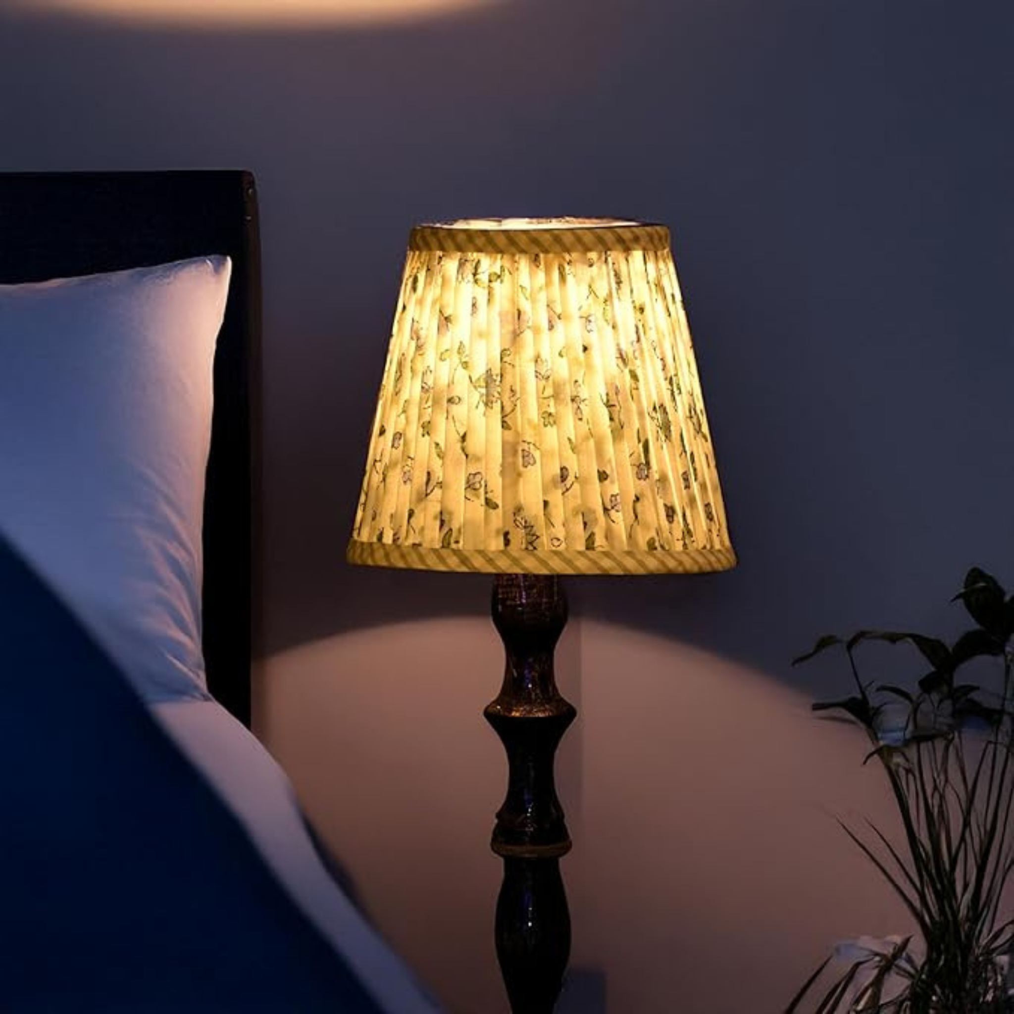 Lamp with a textured shade on a bedside table next to a bed in a dimly lit room.