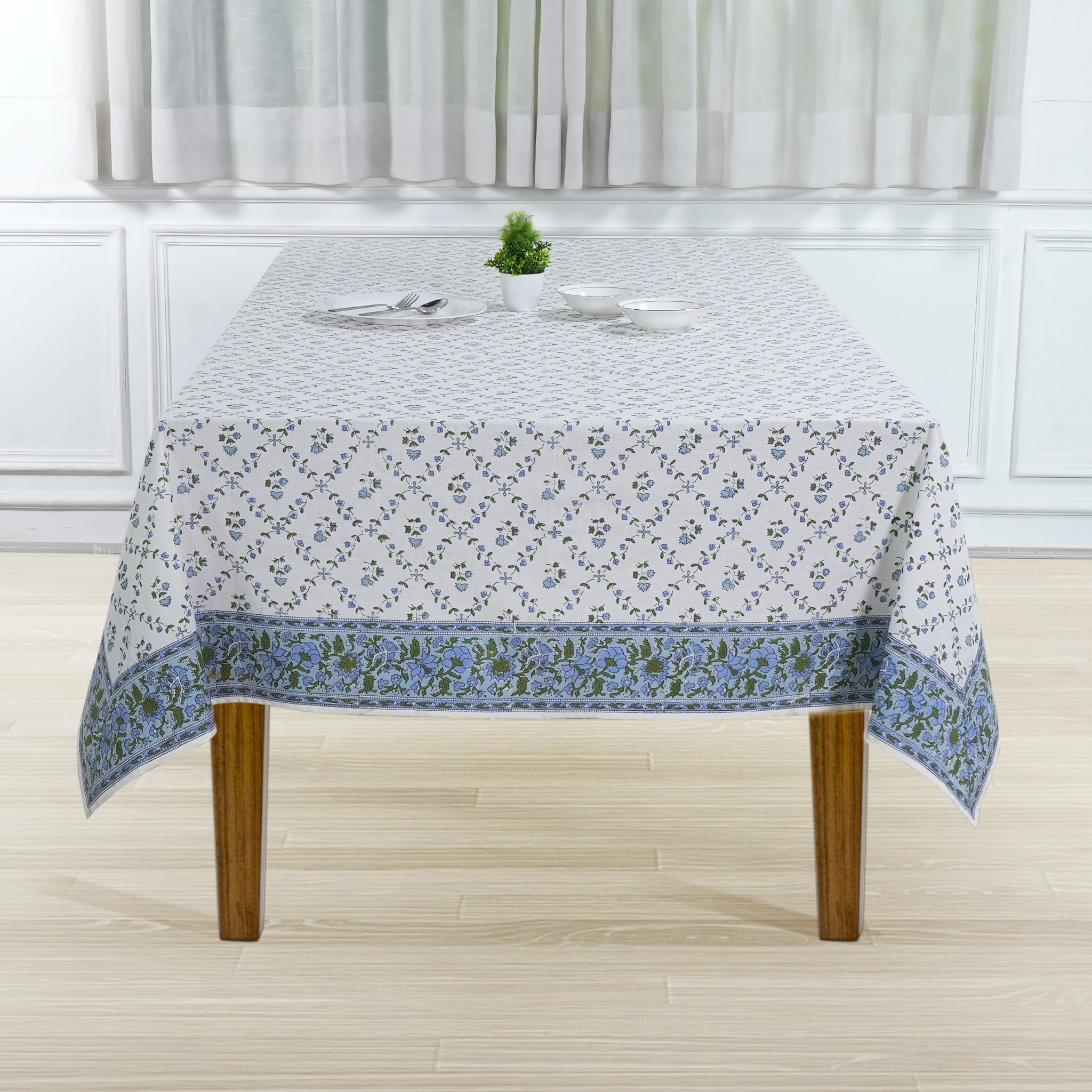 Tablecloth with floral pattern on a wooden table in a room with white walls and curtains.