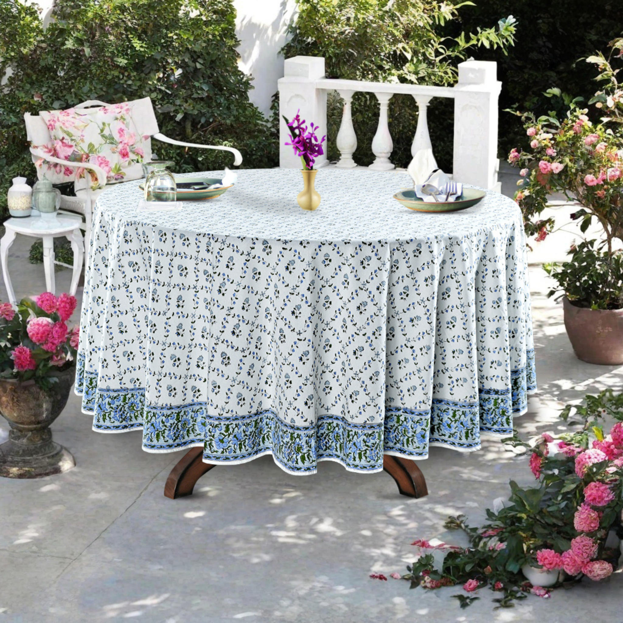 Round table with a floral tablecloth outdoors, surrounded by potted plants.