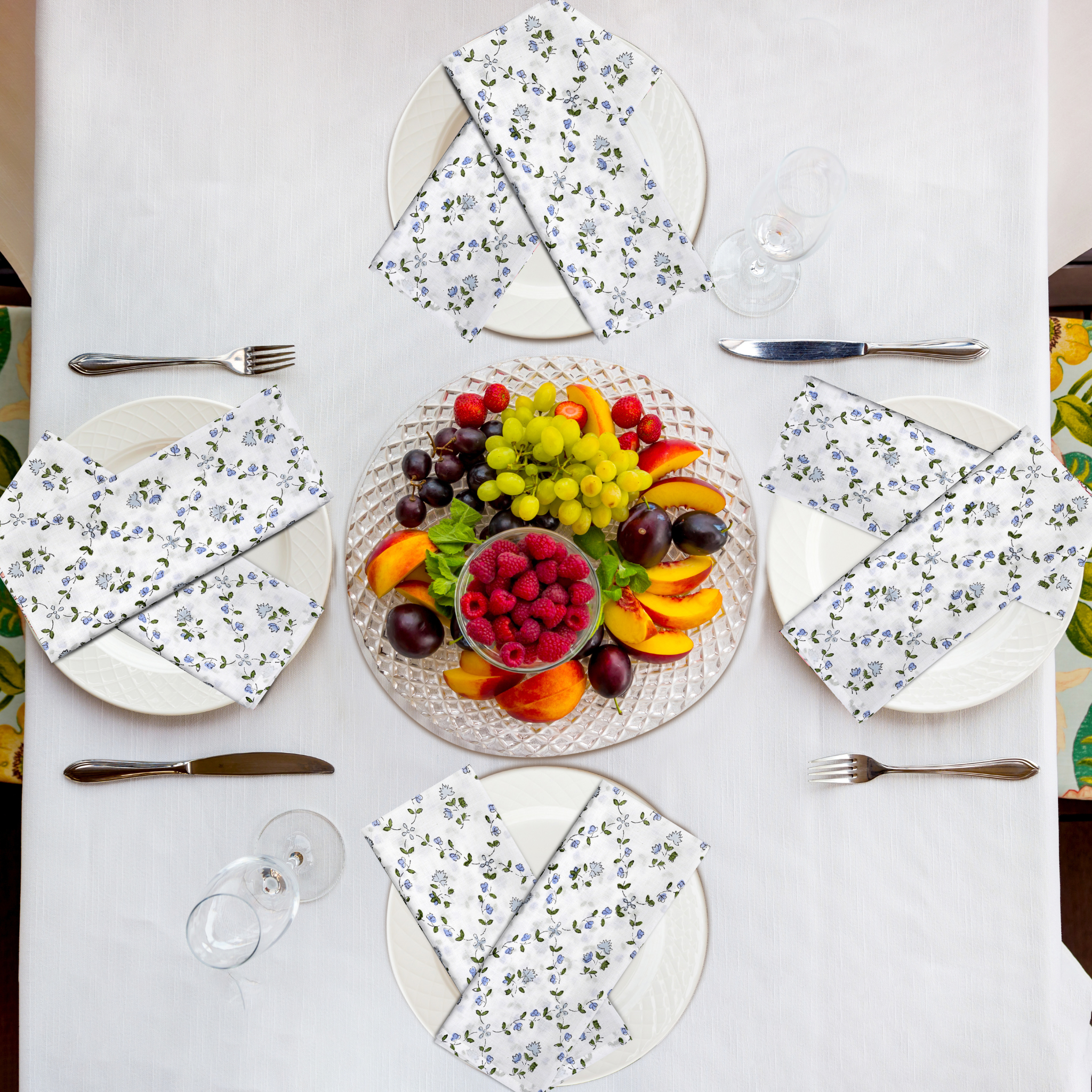 Table setting with floral plates, fruit platter, and cutlery on a white tablecloth.