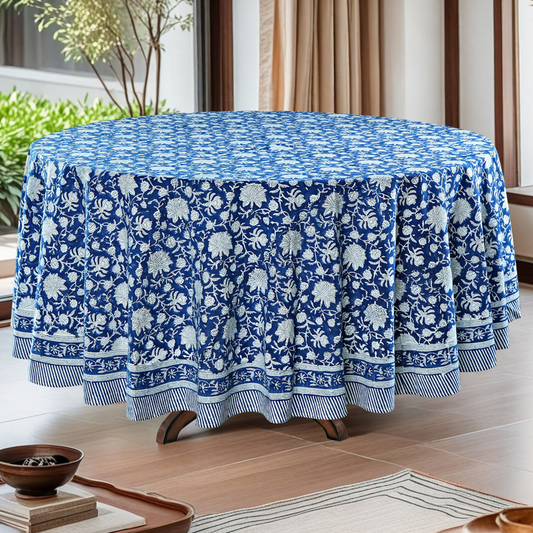 Round table covered with a blue floral patterned tablecloth in a room with plants and curtains.
