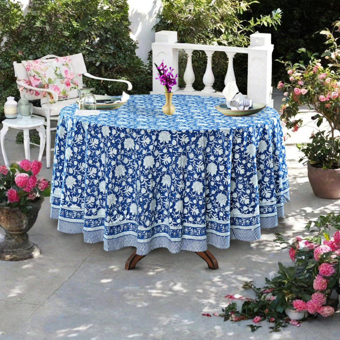 Outdoor setting with a table covered by a blue floral-patterned tablecloth, surrounded by potted plants.