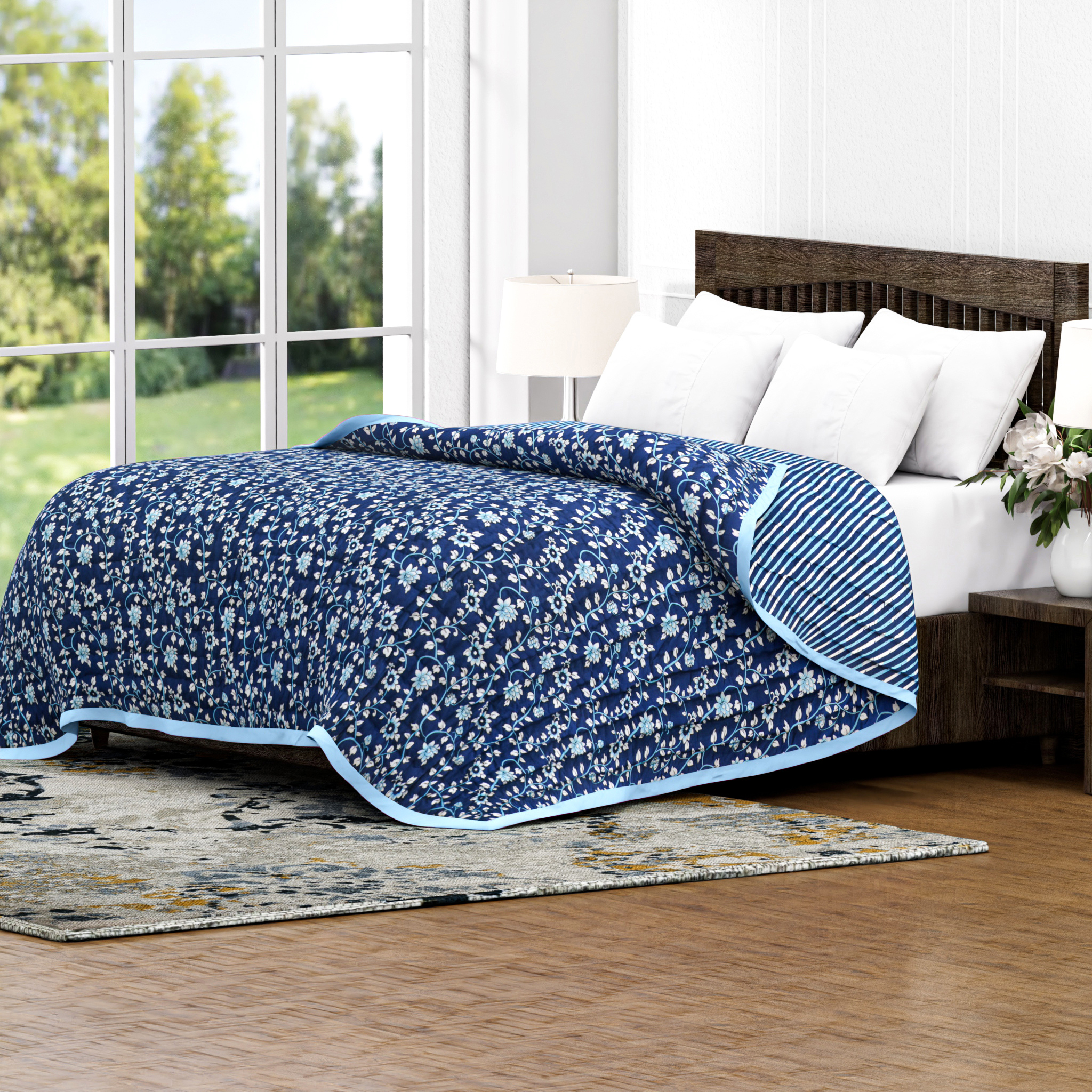 Blue floral quilt on a bed in a room with large windows and greenery outside.