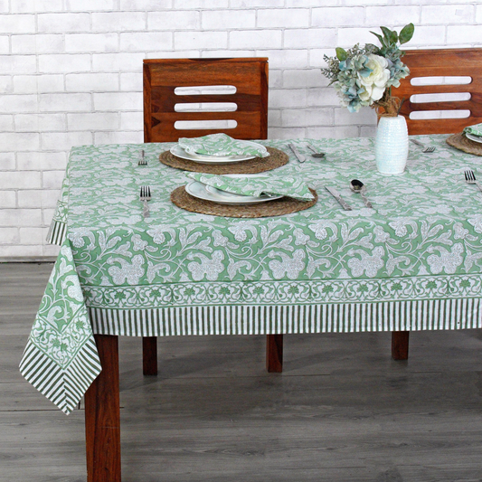 Dining table set with a green and white patterned tablecloth, wooden chairs, and a vase with flowers.