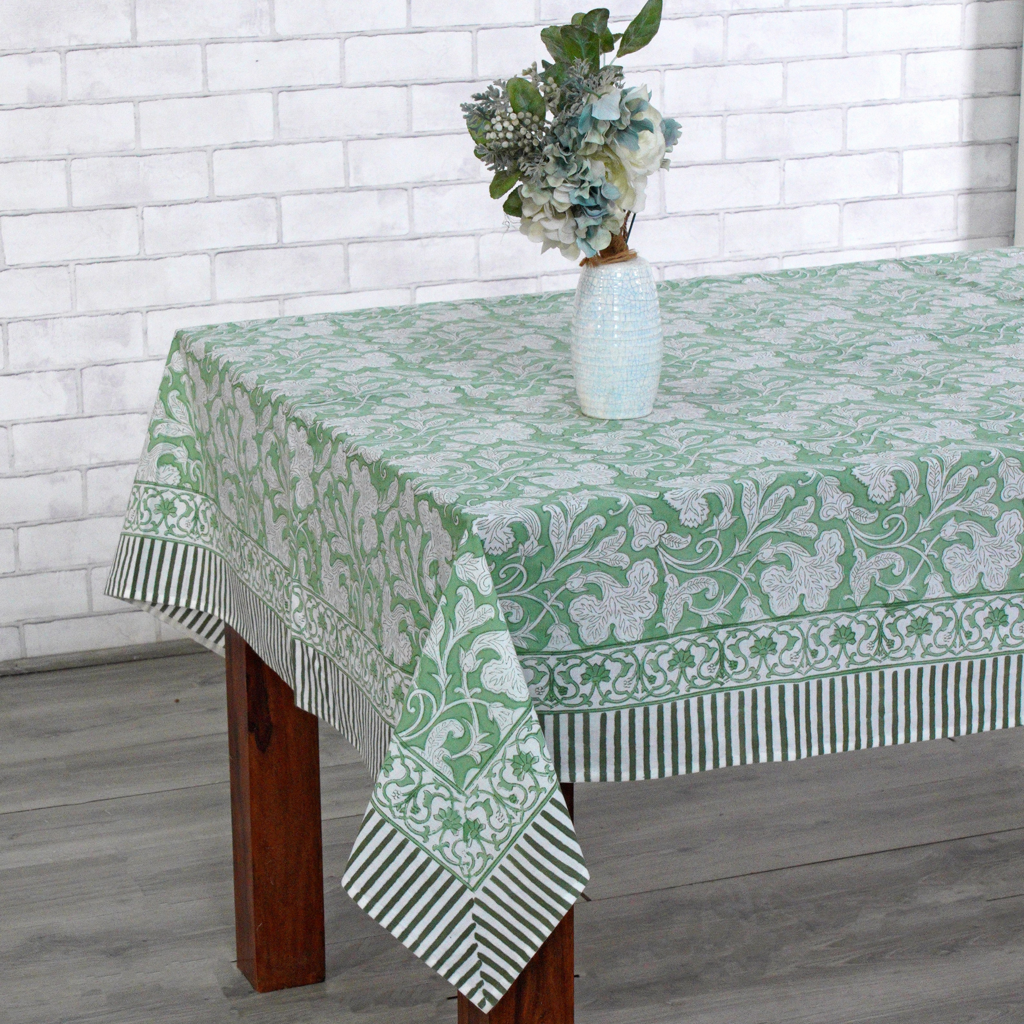 Tablecloth with green floral pattern on a wooden table against a white brick wall.