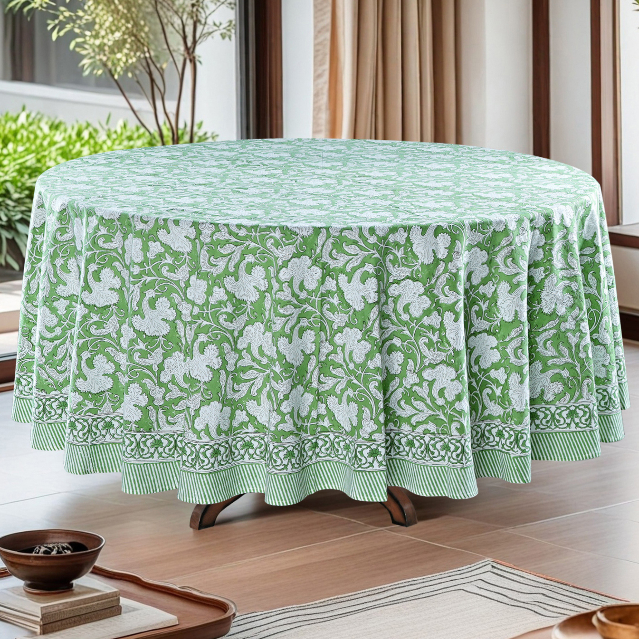 Green floral patterned tablecloth on a round table in a room with plants and curtains.