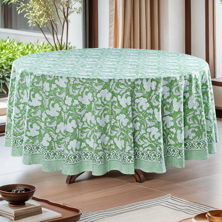 Green floral patterned tablecloth on a round table in a room with plants and curtains.