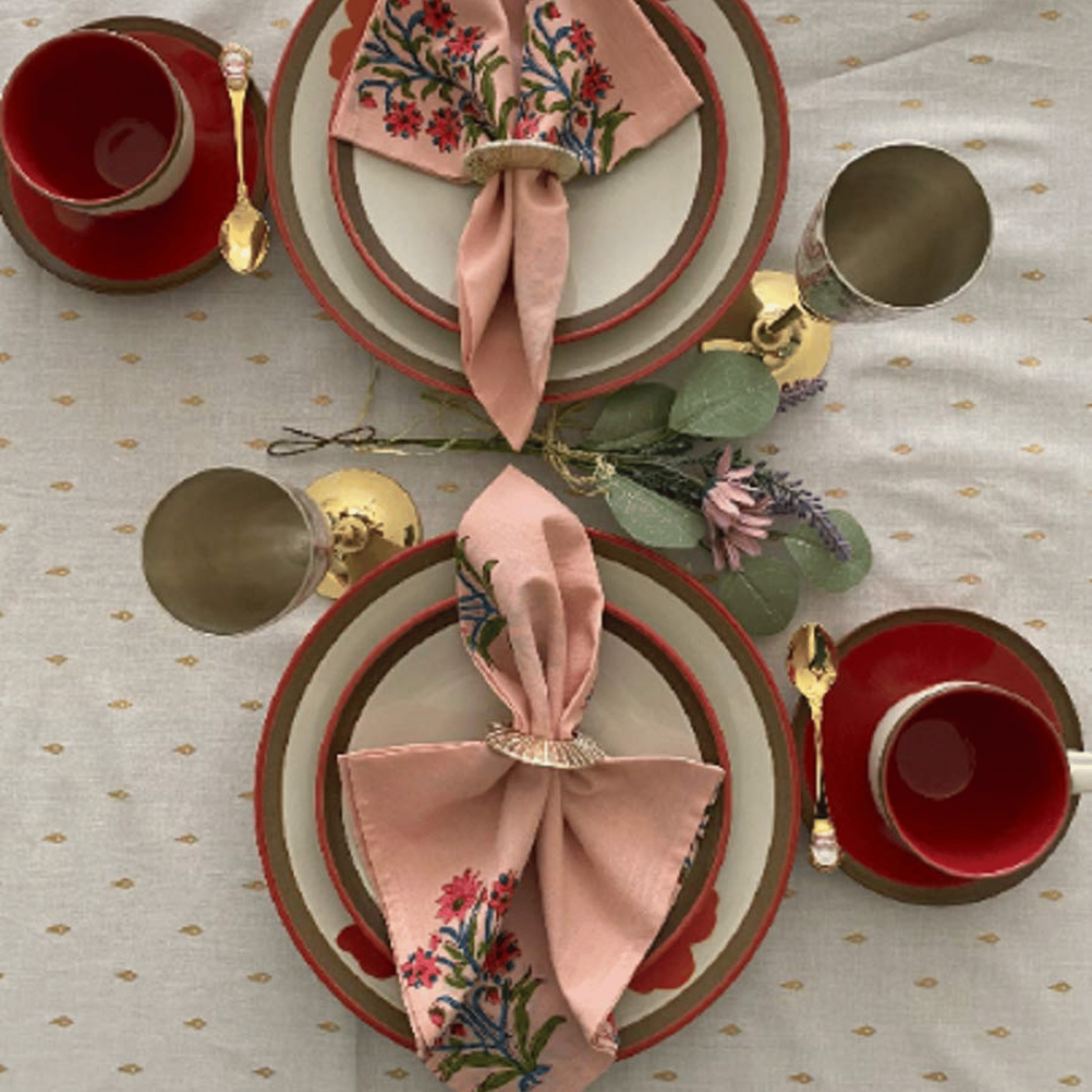 Elegant table setting with red cups, gold saucers, and floral napkins on a patterned tablecloth.