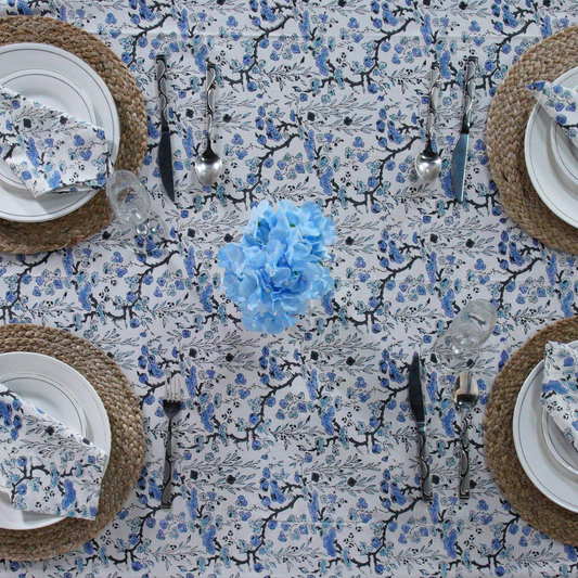 Table setting with floral tablecloth, plates, cutlery, and a blue flower centerpiece.
