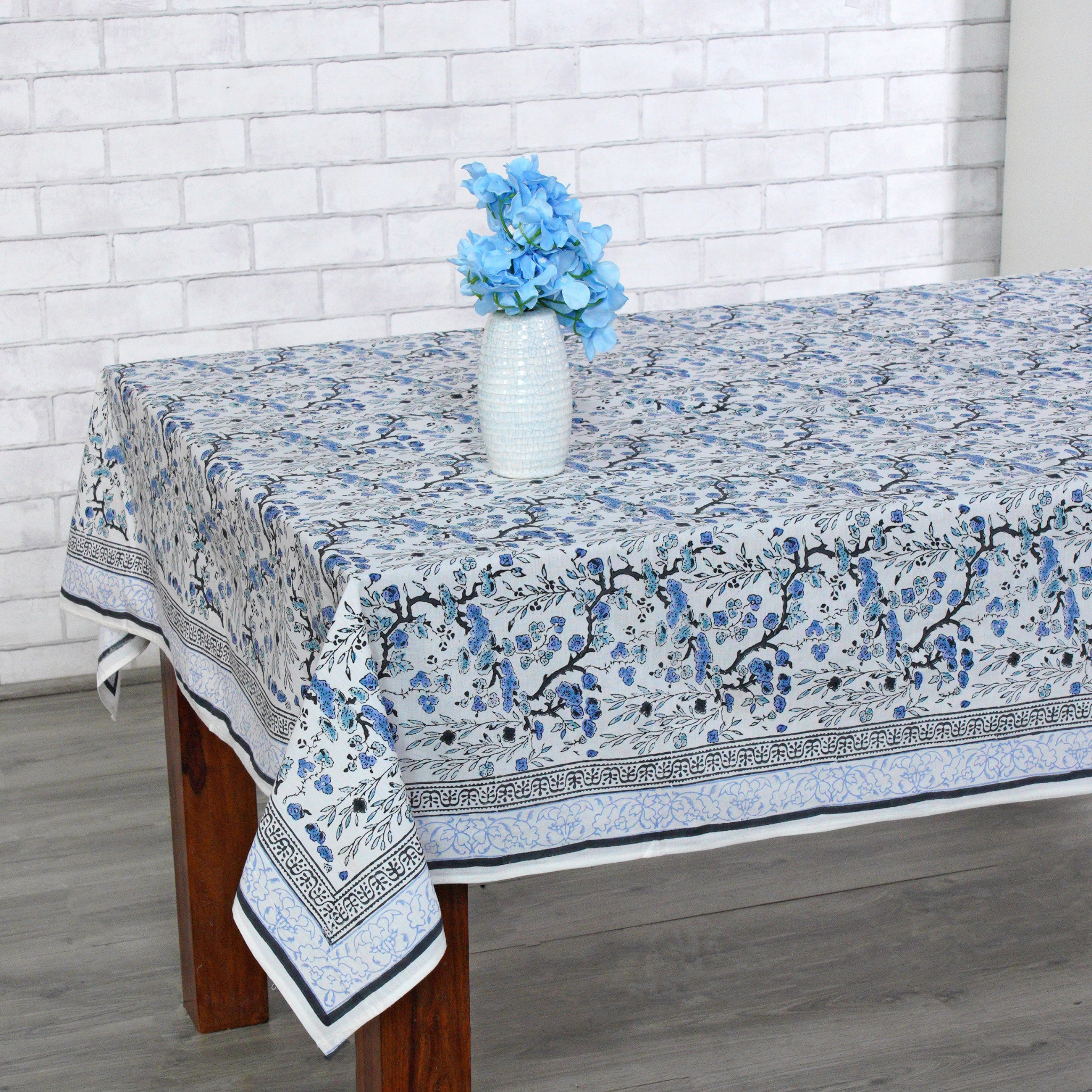 Tablecloth with floral pattern on a wooden table against a white brick wall.