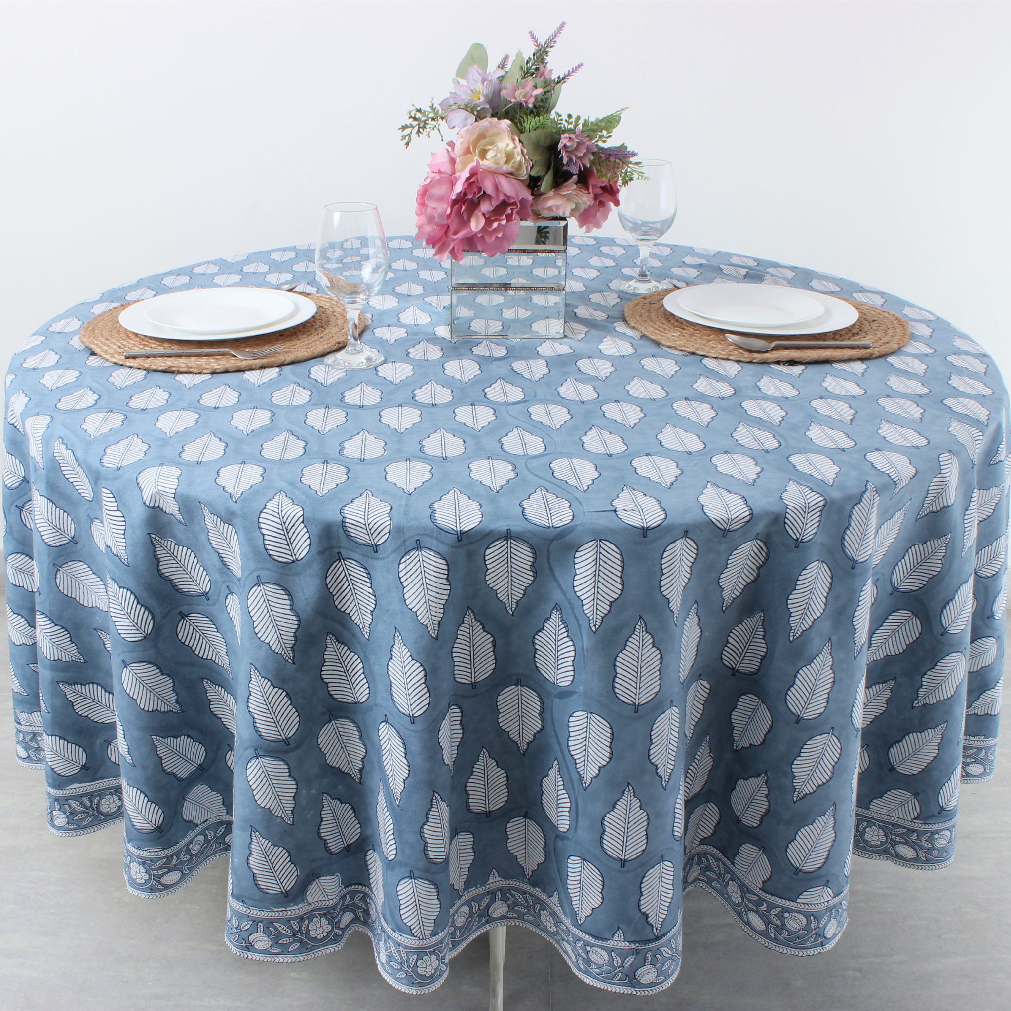 Round table with a blue patterned tablecloth, white plates, and floral centerpiece on a light background