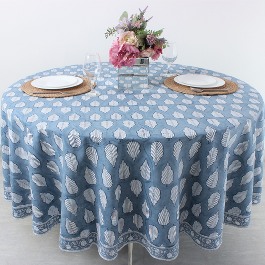 Round table with a blue patterned tablecloth, white plates, and floral centerpiece on a light background