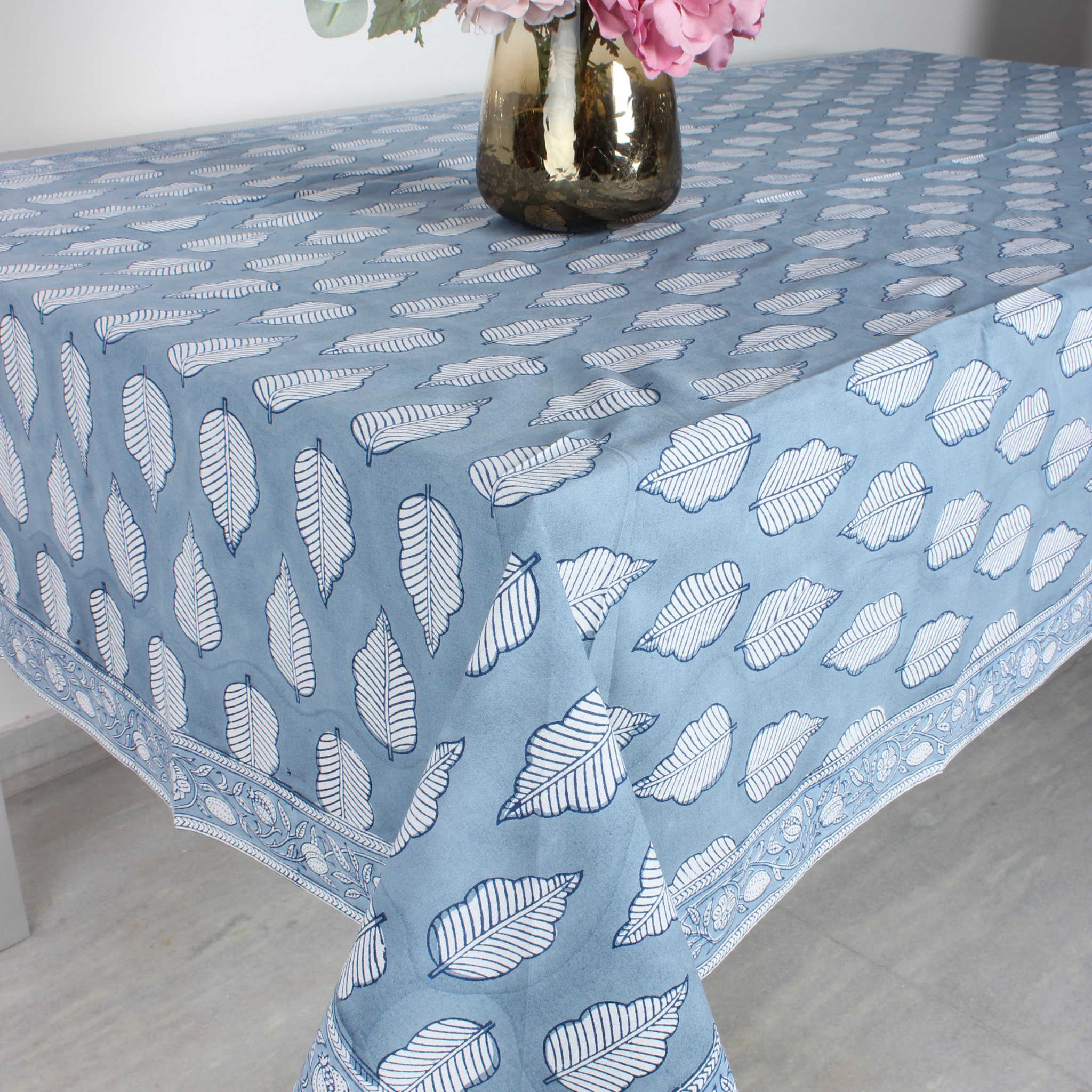 Blue tablecloth with white leaf pattern on a table, with a vase of flowers in the background.