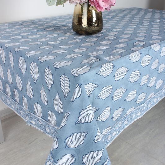 Blue tablecloth with white leaf pattern on a table, with a vase of flowers in the background.
