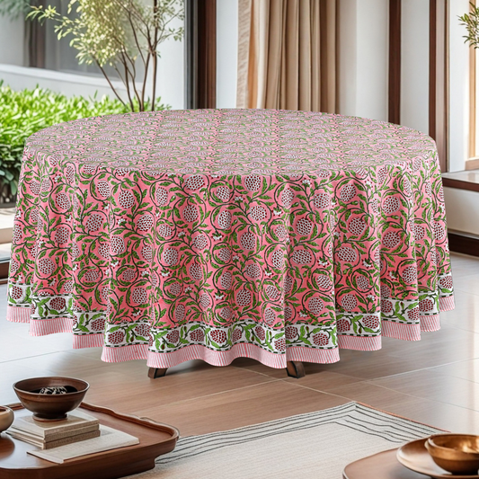 Round table with a pink and green patterned tablecloth in a room with plants and curtains.