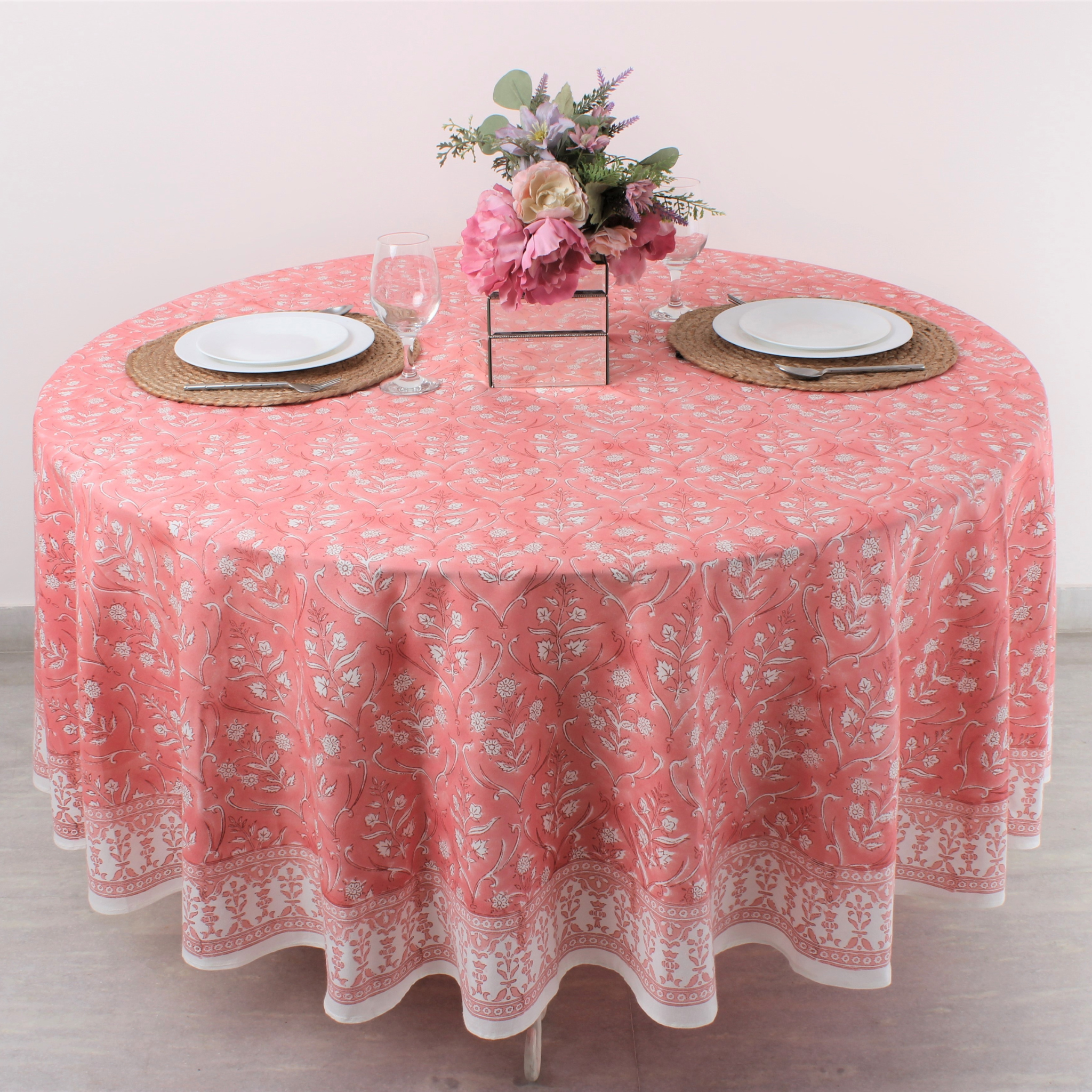 Round table with a pink floral tablecloth, plates, and flowers on a white background
