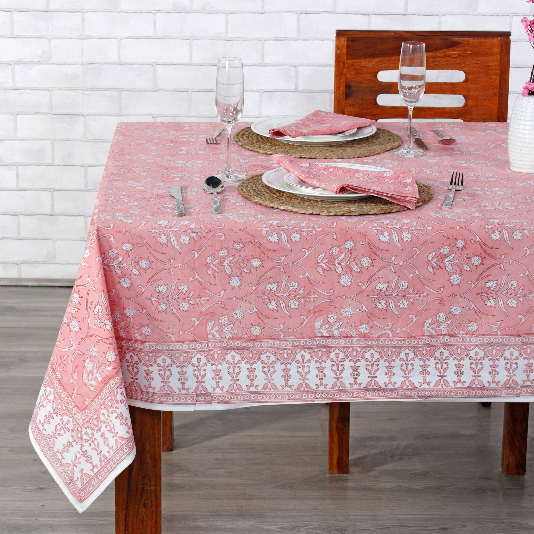 Dining table set with a pink patterned tablecloth against a white brick wall.
