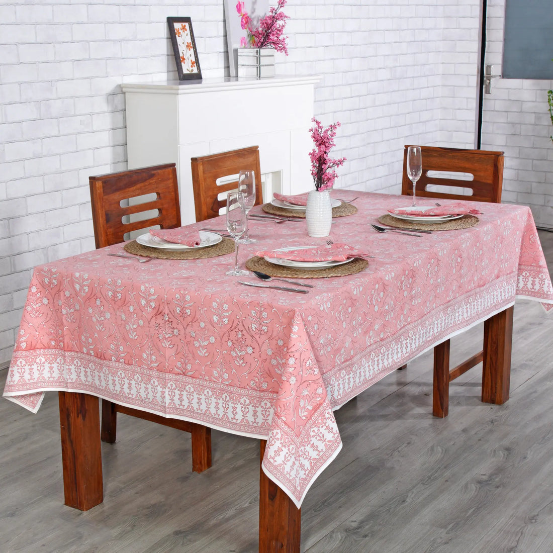 Dining table set with a pink tablecloth and wooden chairs against a white brick wall.