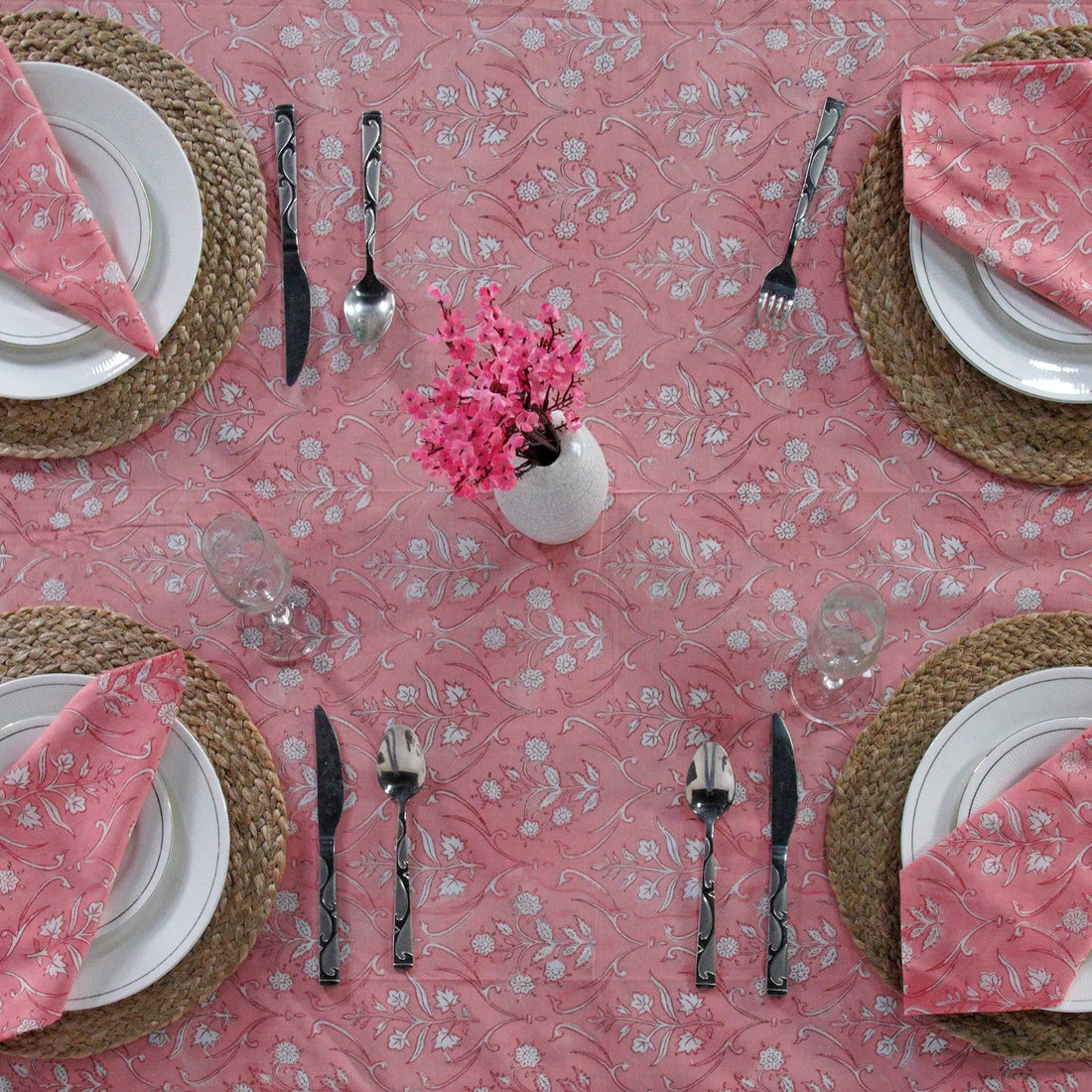 Dining table setting with pink floral tablecloth, plates, cutlery, and napkins.
